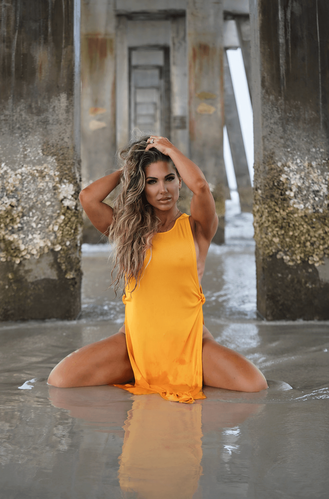 Woman in yellow dress kneeling in water under a bridge, with hands in her hair, looking at the camera.