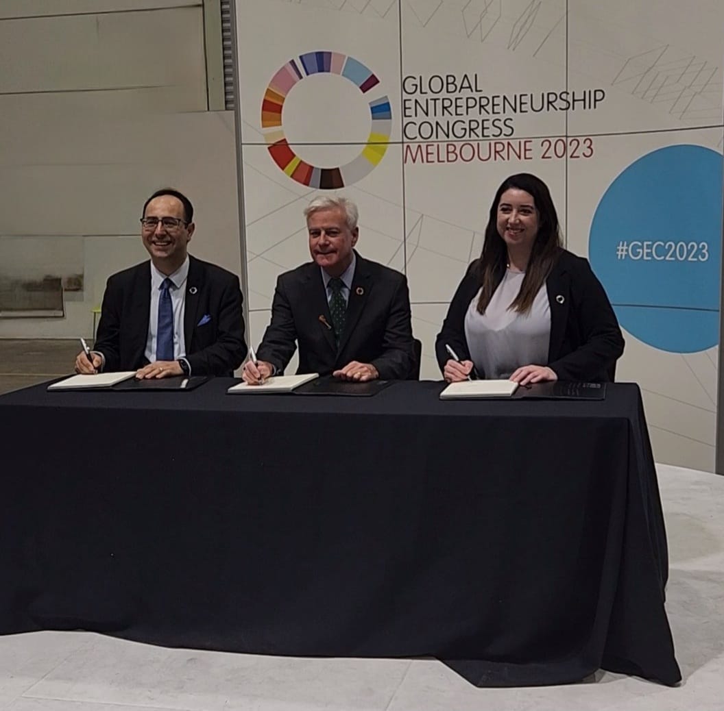 Three people sitting at a table signing documents at the Global Entrepreneurship Congress Melbourne 2023, with a banner behind them.