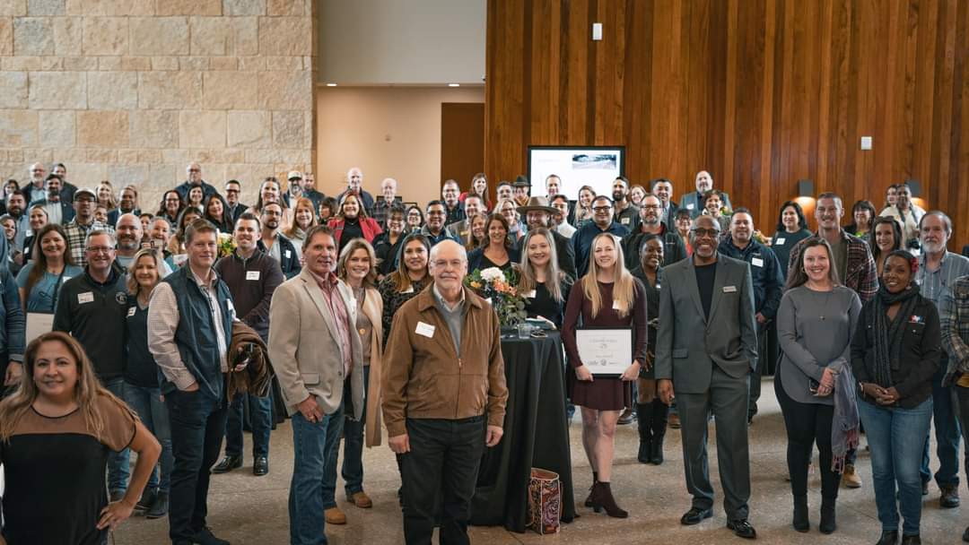 Group of diverse people gathered indoors, some holding certificates, in a spacious room with wooden and stone walls.