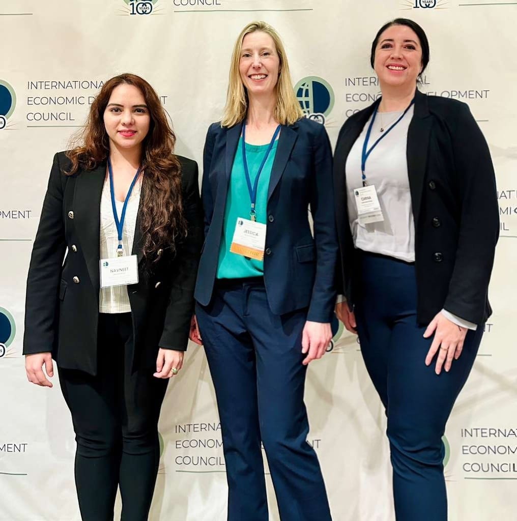 Three women in business attire standing in front of a backdrop with logos, smiling at the camera.