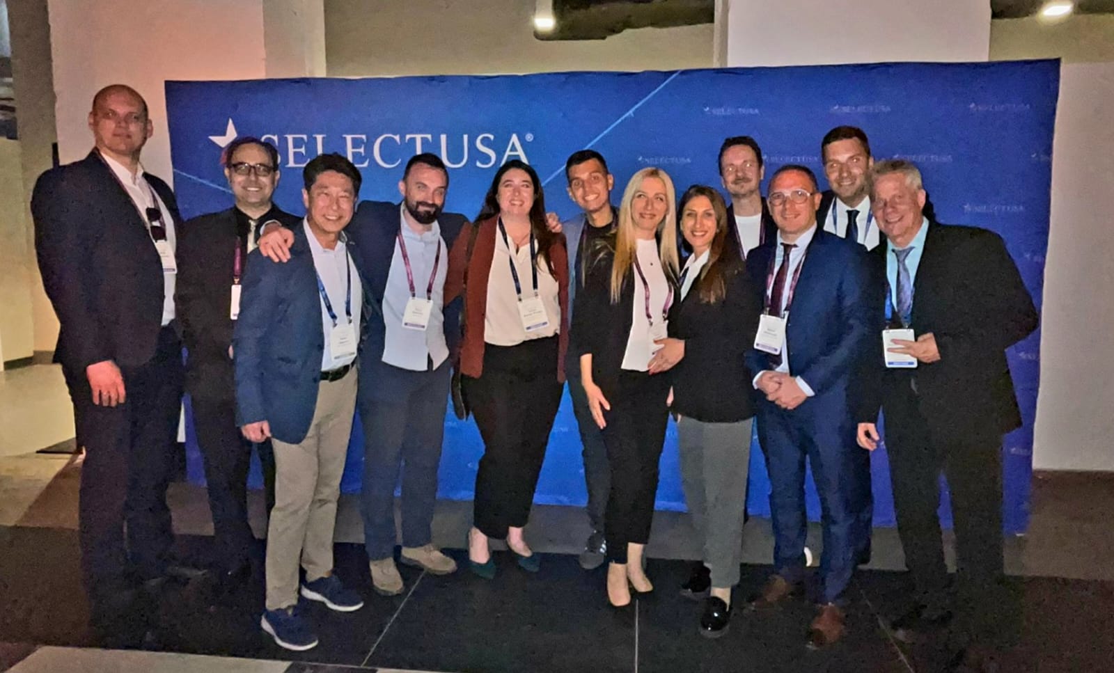 Group of eleven people standing in front of a blue banner with white text, posing for a photo at an event.