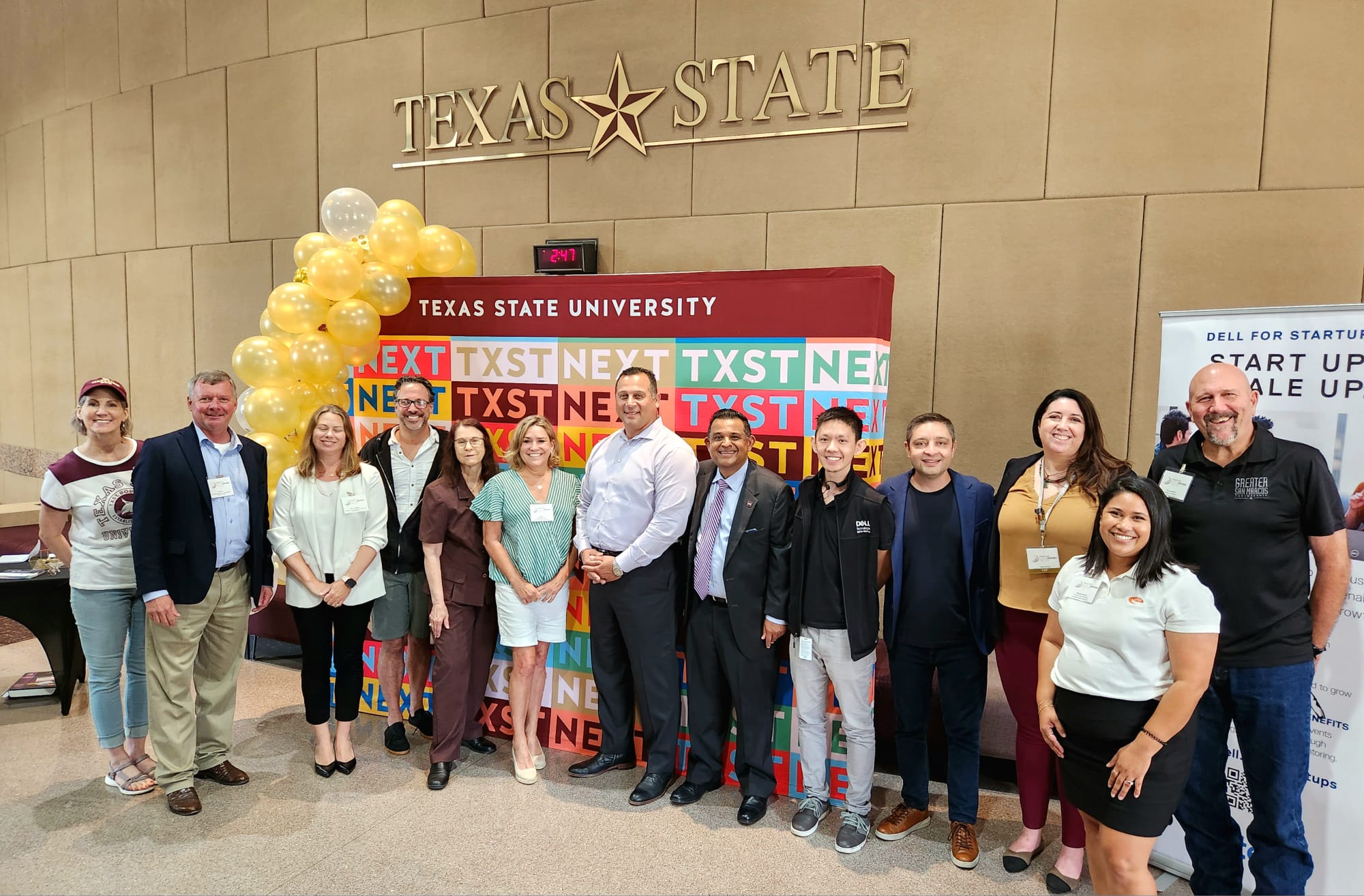 Group of people standing in front of a Texas State University sign with balloons and banners, indoors.