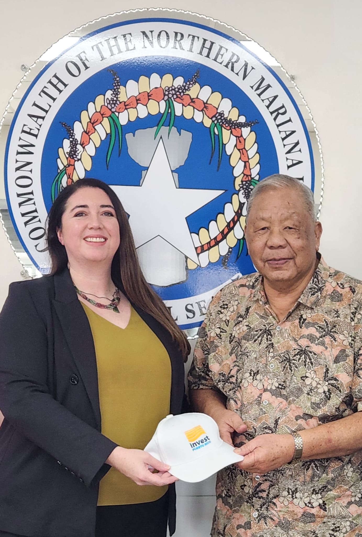 Two people standing in front of a seal, one holding a white cap, smiling. Seal reads 'Commonwealth of the Northern Mariana Islands'.