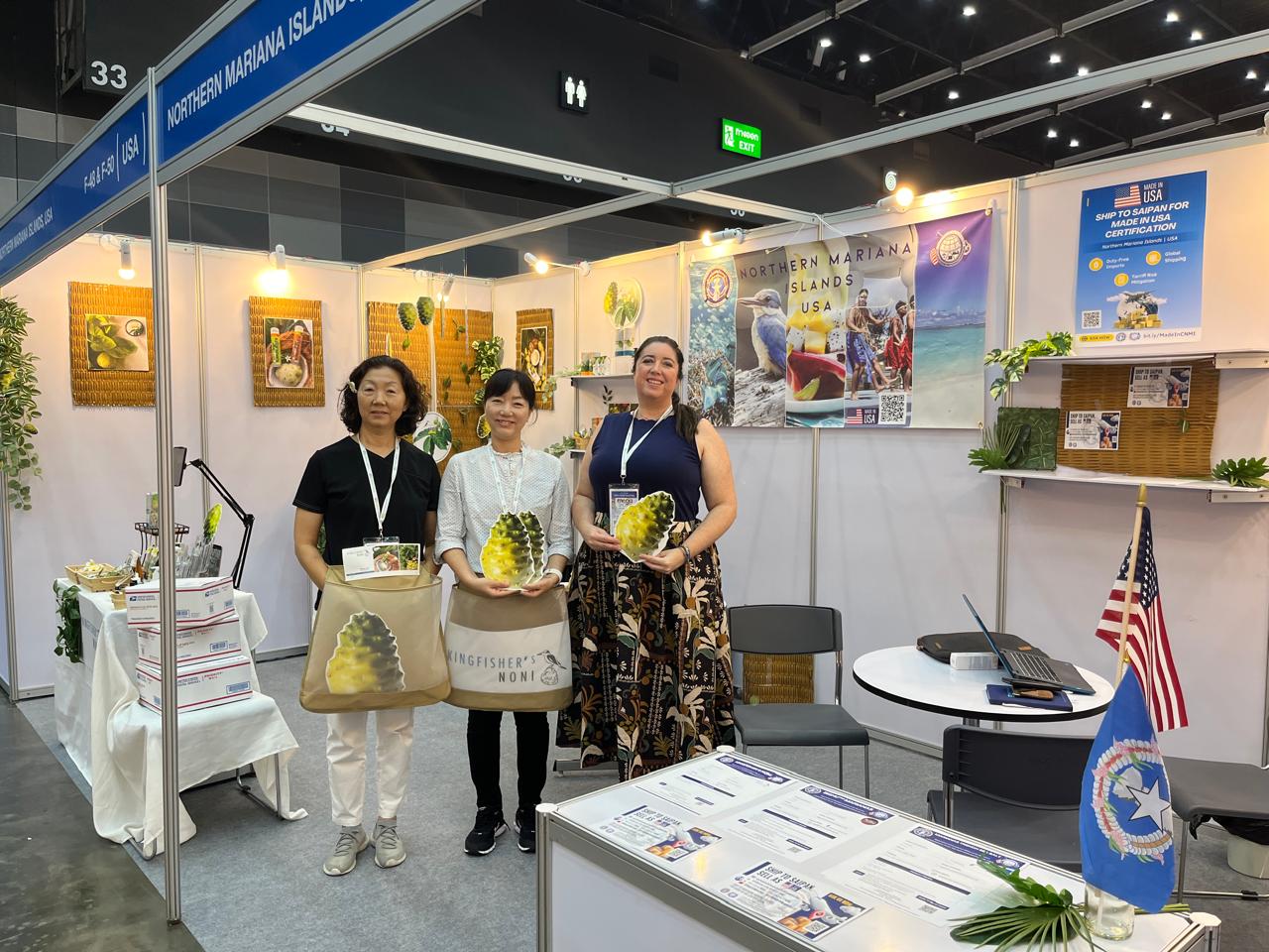 Three women stand in an exhibition booth with posters and displays, holding pineapple-shaped objects, with a table and American flag nearby.