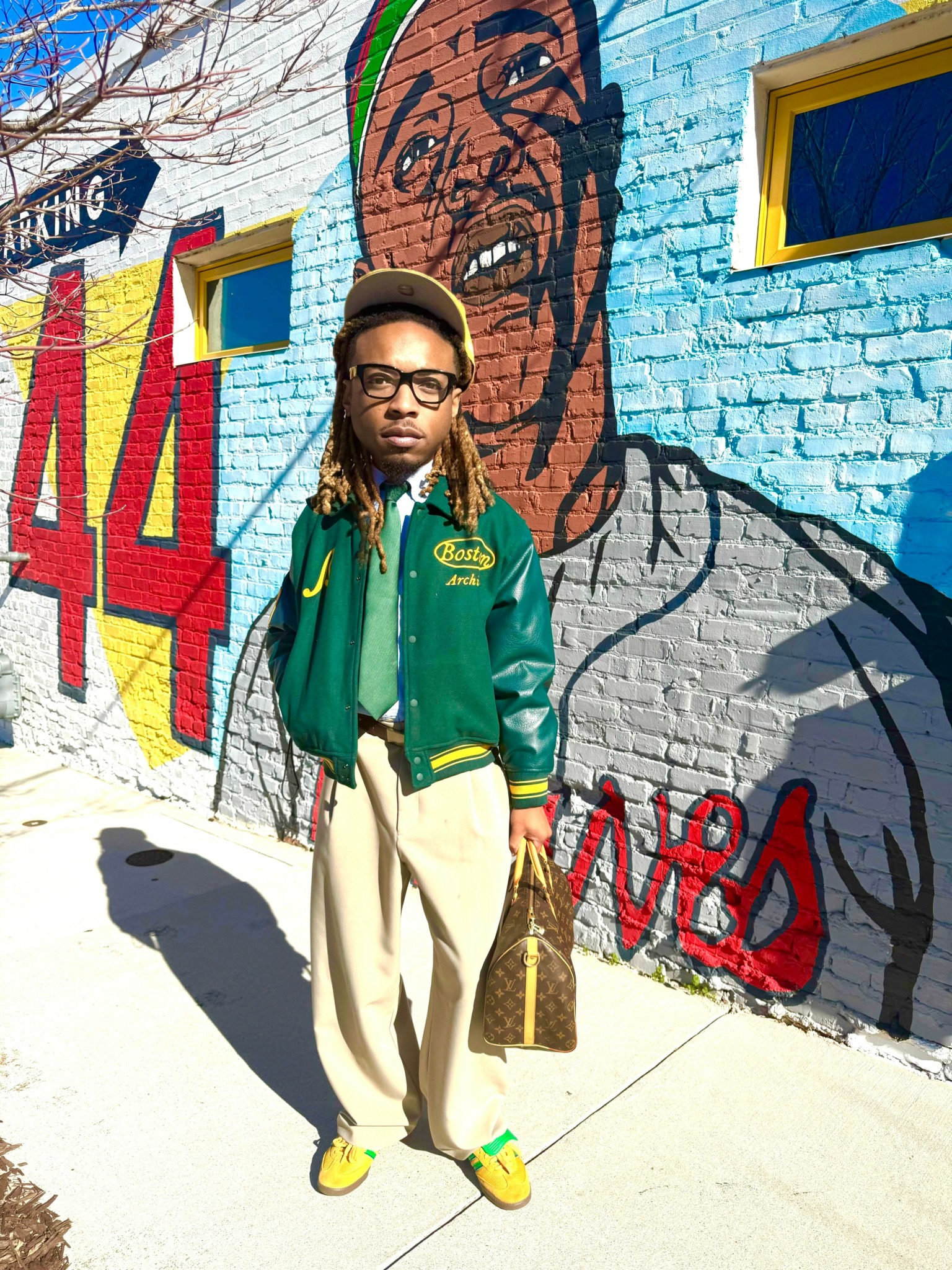 Person standing in front of a colorful mural on a brick wall, wearing glasses, a green jacket, and beige pants.