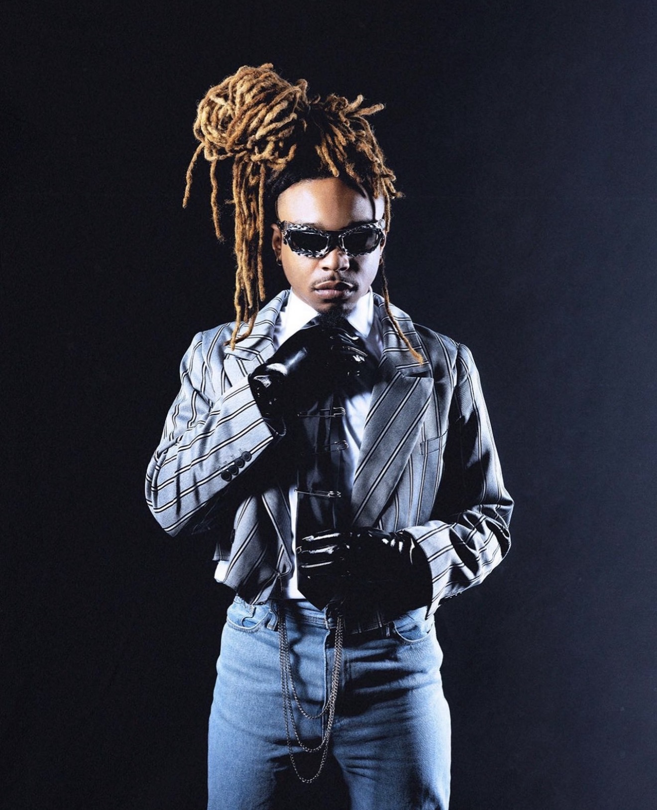 Person with dreadlocks wearing sunglasses, a striped jacket, gloves, and holding a camera, standing against a dark background.