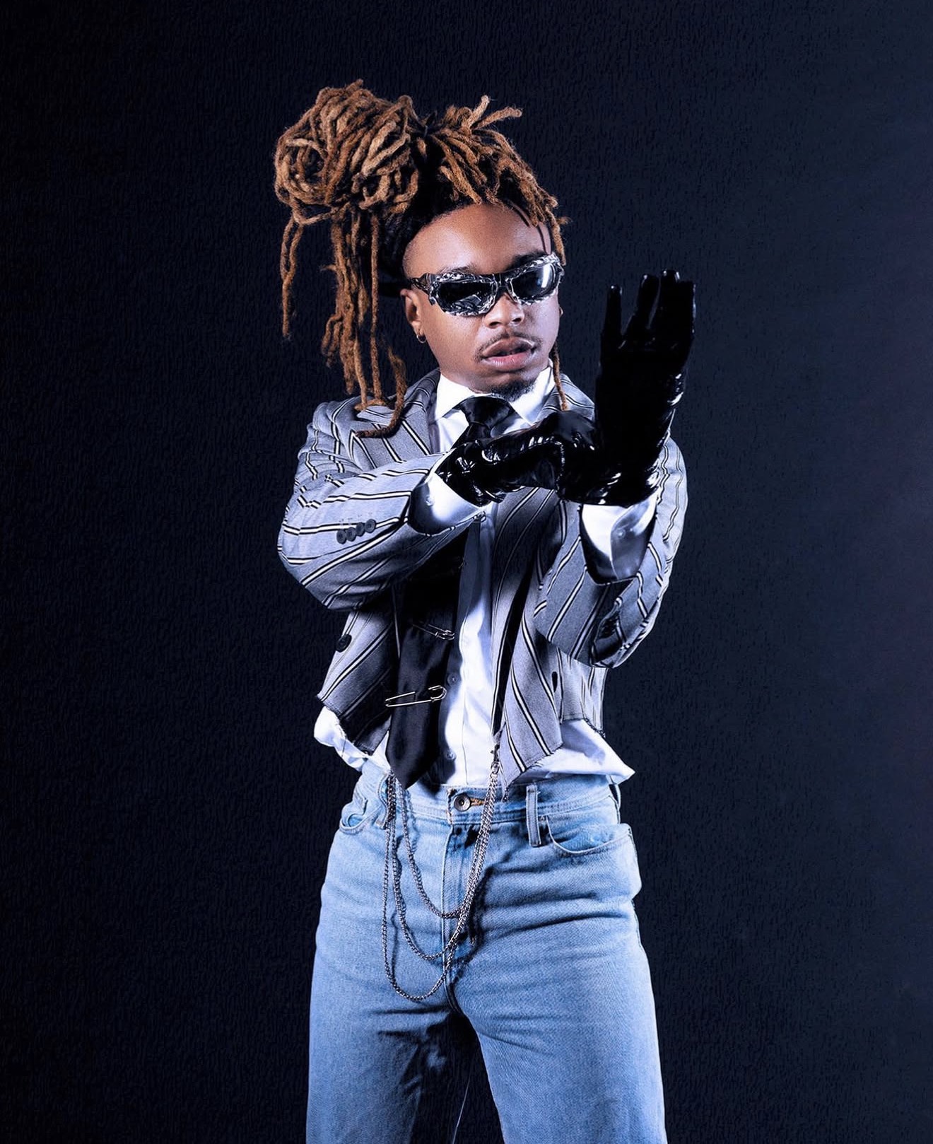 Person with dreadlocks wearing sunglasses, a striped jacket, gloves, and jeans, posing against a dark background.