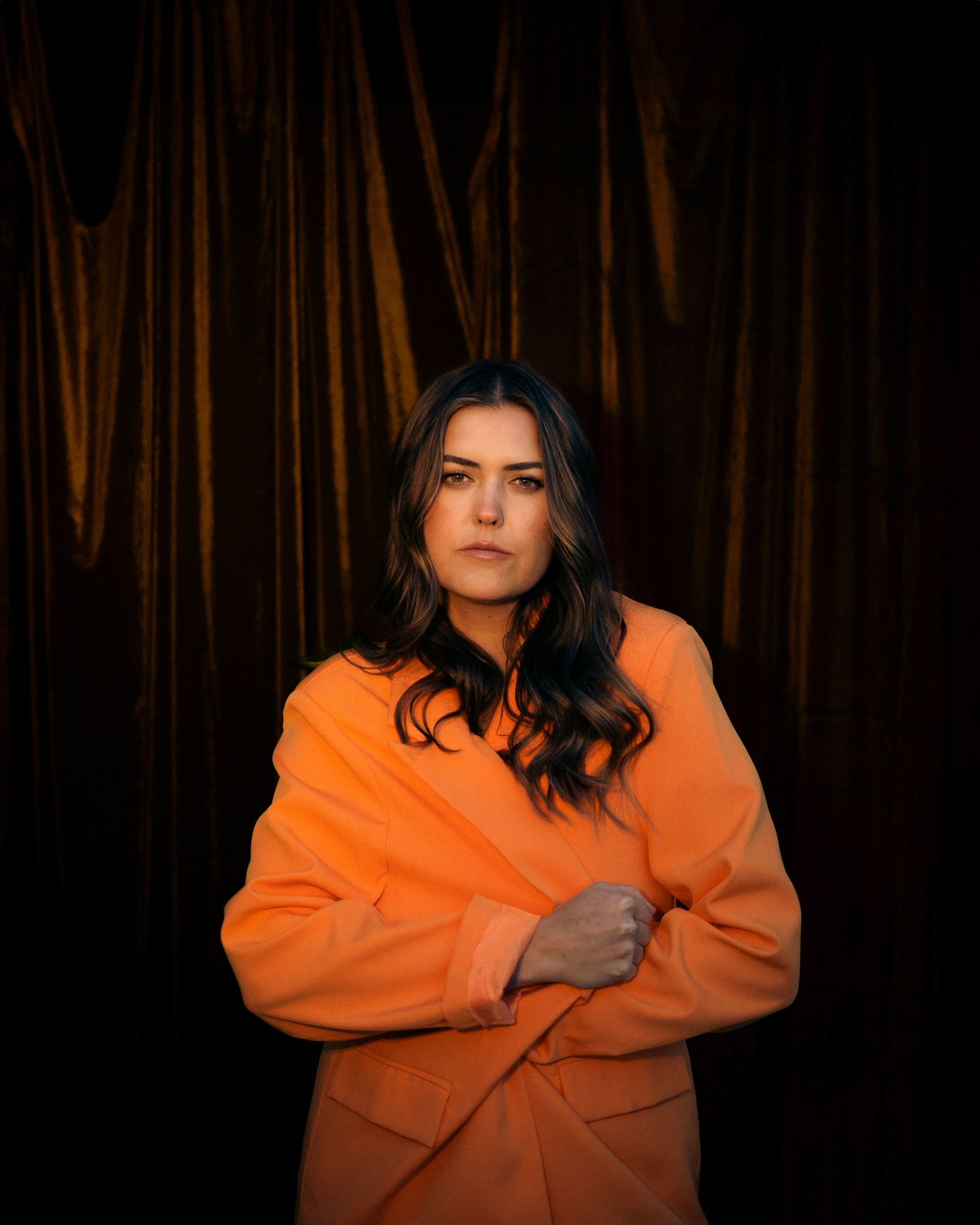 Woman with long dark hair wearing an orange coat, standing in front of dark curtains.