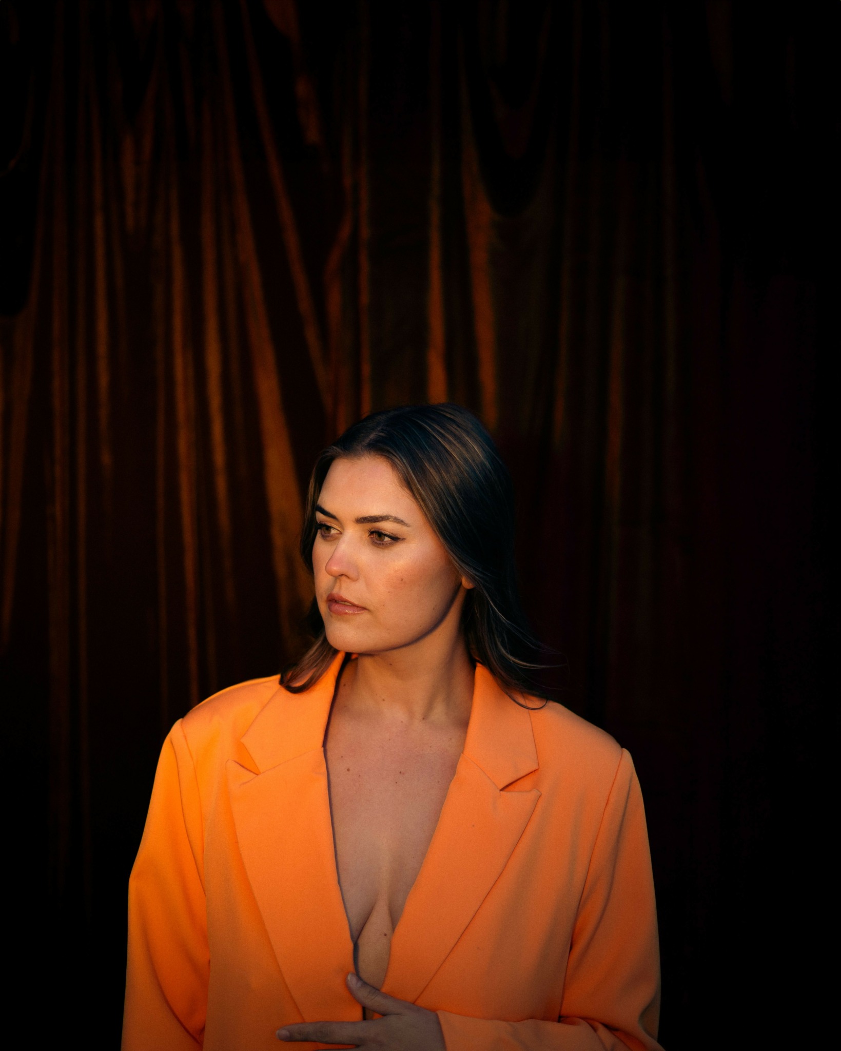 Woman with dark hair in an orange blazer, standing against a dark curtain background.