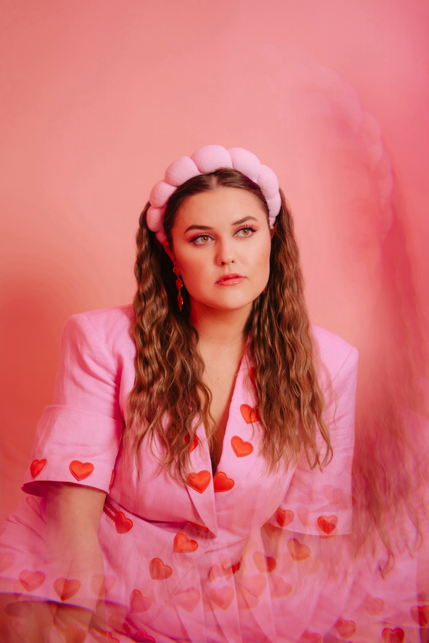 Young woman with long wavy hair wearing a pink blazer with heart patterns and a pink headband, against a pink background.