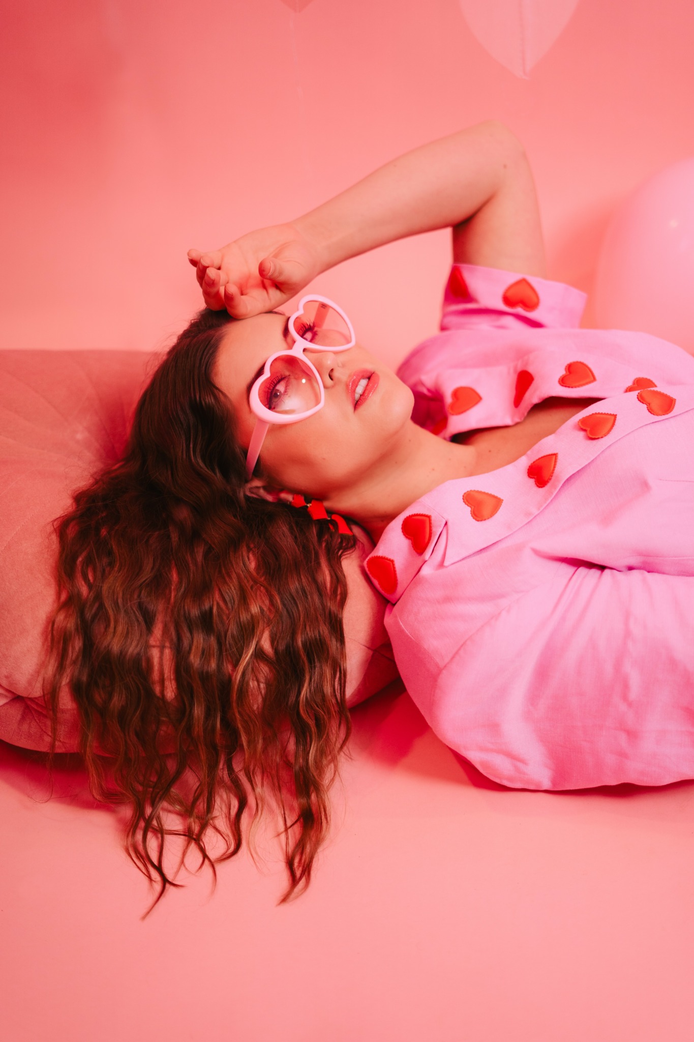 Woman with curly hair lying on pink background, wearing heart-shaped sunglasses and a pink dress with red hearts.