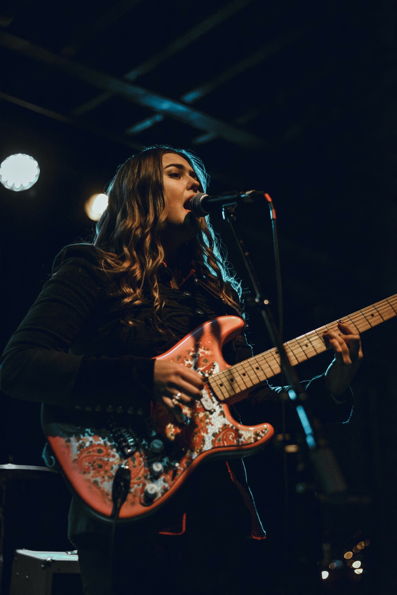 Female musician singing into microphone while playing an electric guitar on stage, with stage lights overhead.