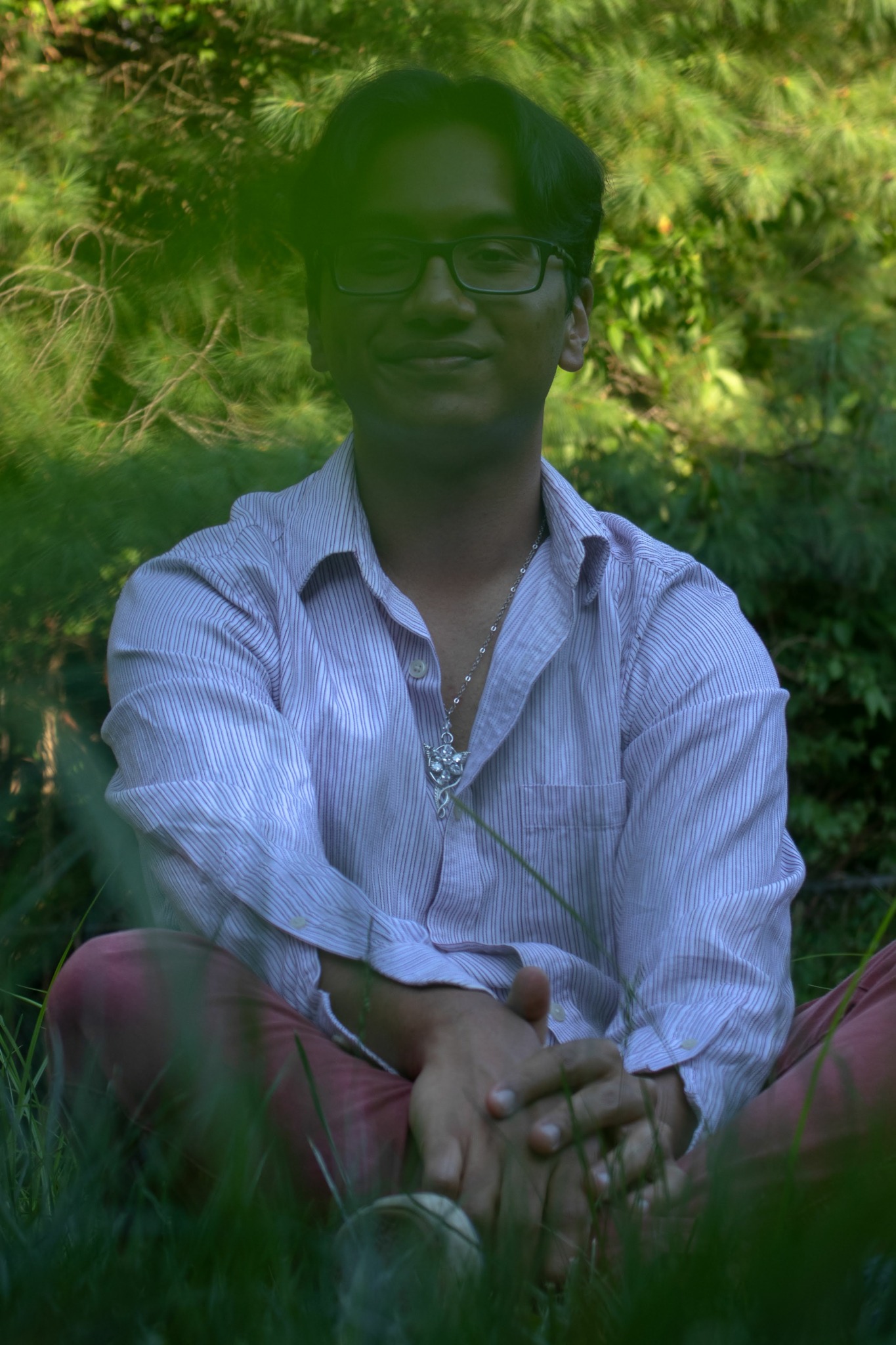 Person sitting outdoors with trees and greenery in background, wearing glasses and a light-colored shirt, hands clasped.