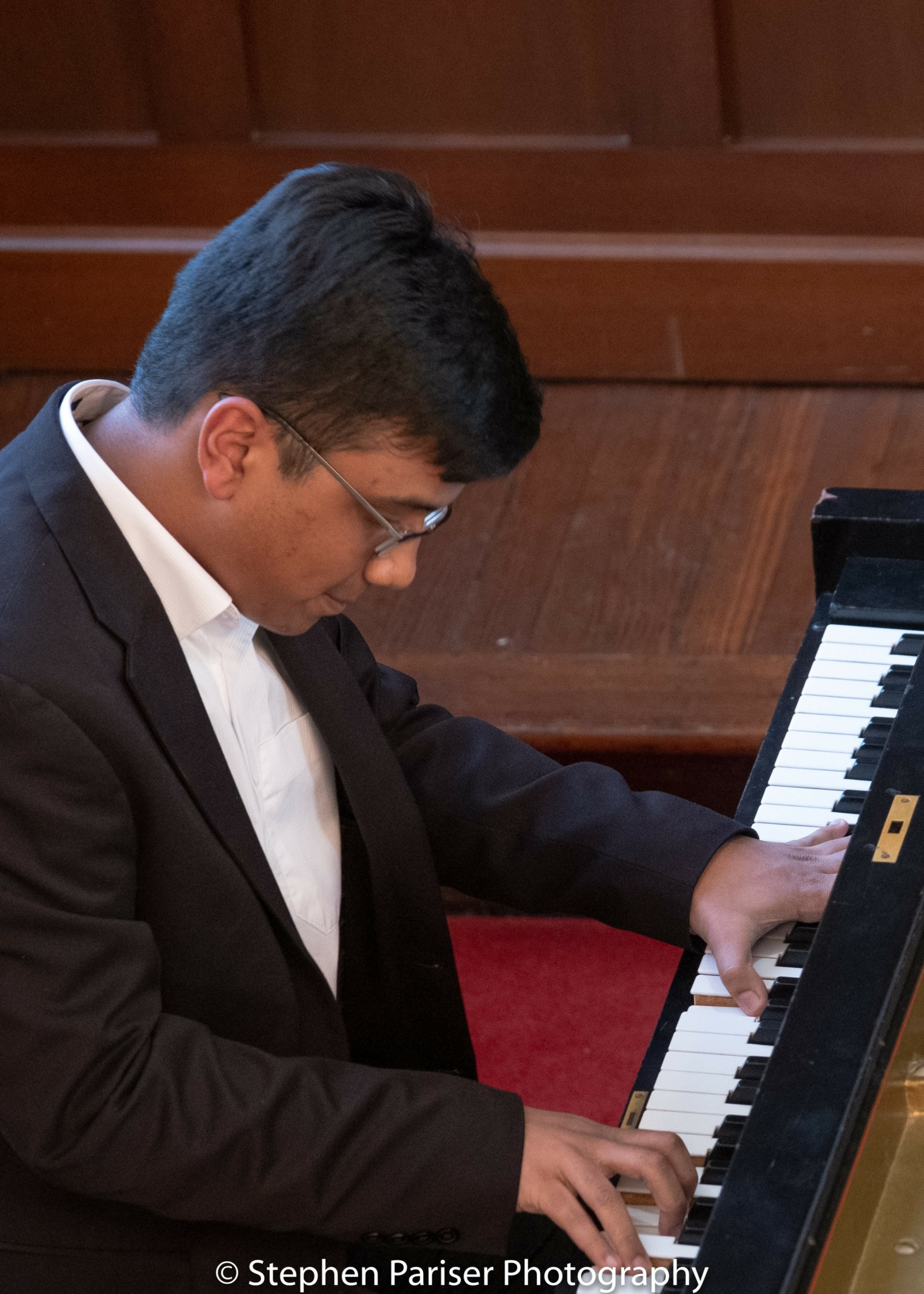Person playing piano, wearing glasses and a suit, in a wooden room, focused on the keys.