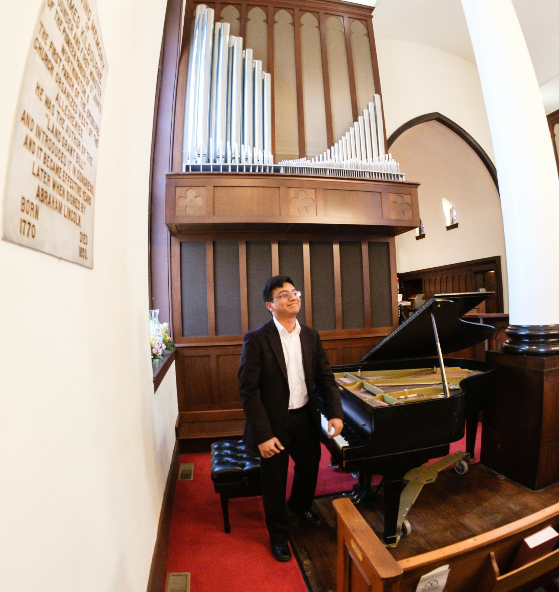 Man in suit standing next to a grand piano in a church with pipe organ and wooden architecture.