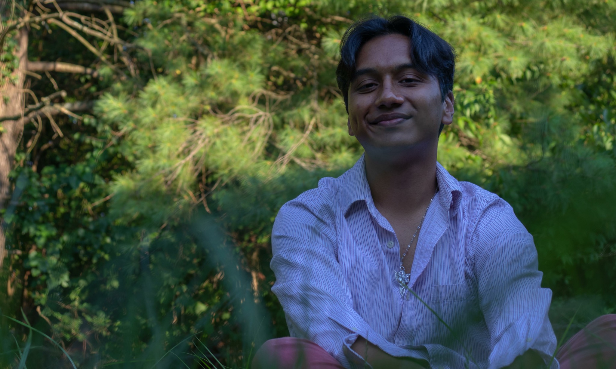 Person with dark hair smiling outdoors among green trees and foliage.
