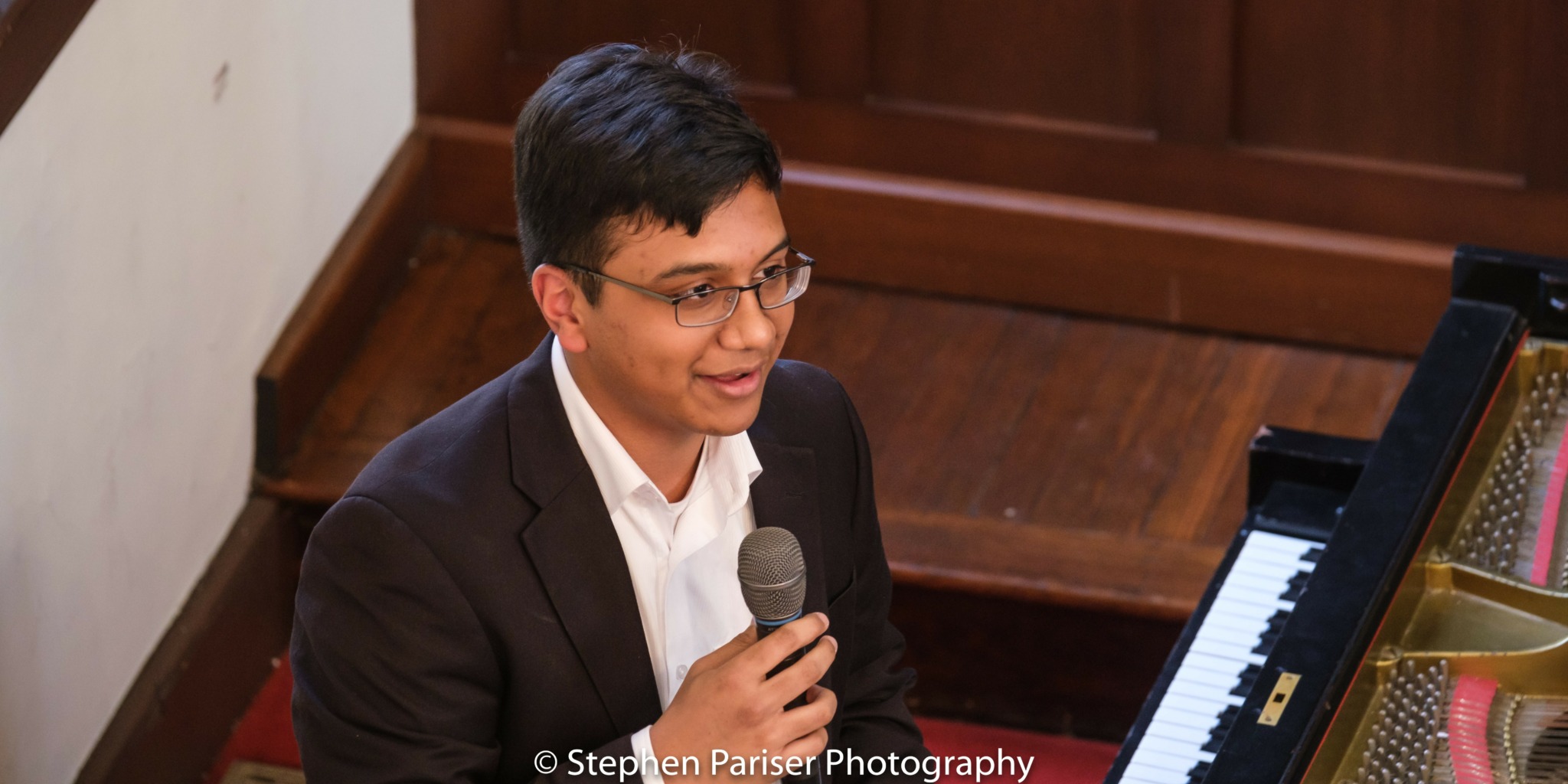 Person with glasses in a suit speaking into a microphone, seated near a piano, in a room with wooden flooring.