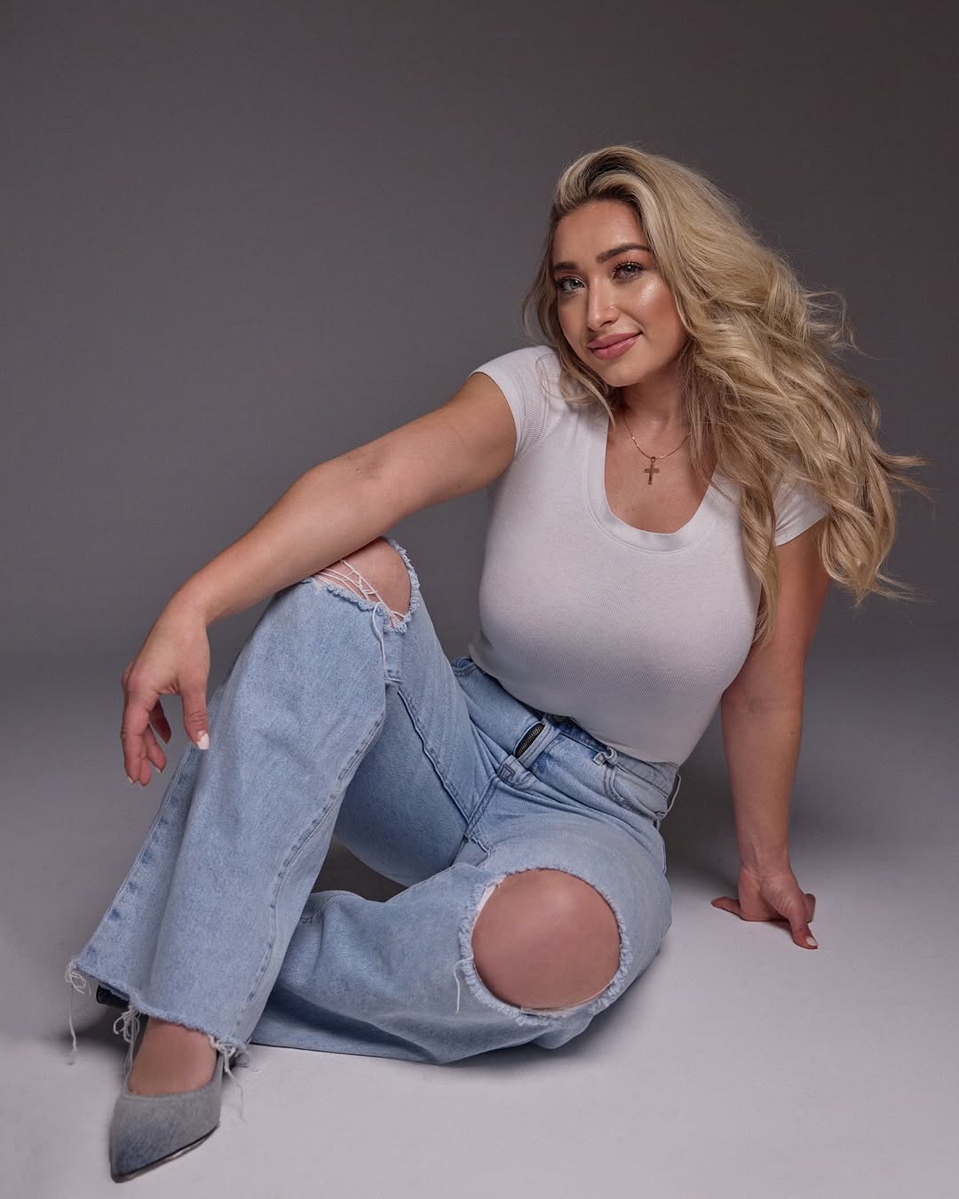 Woman with long blonde hair sitting on the floor, wearing a white t-shirt and ripped jeans, smiling.