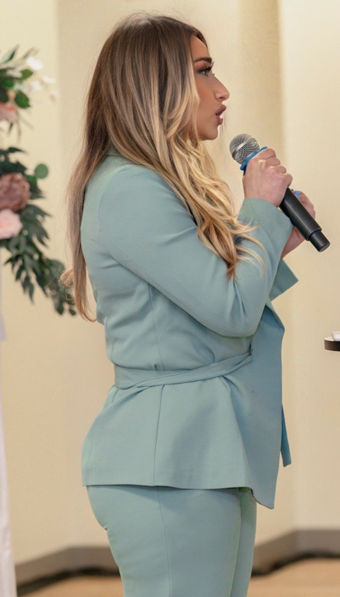 Woman with long hair holding a microphone, wearing a light blue suit, standing indoors with flowers in the background.