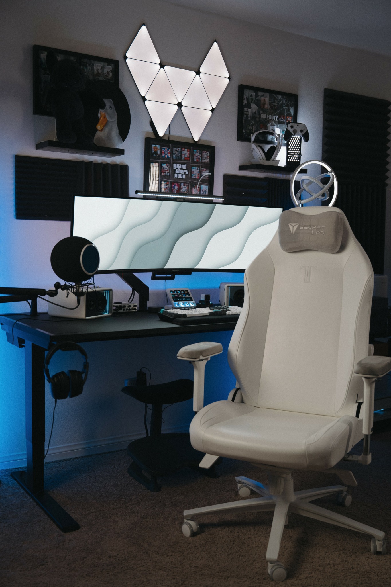 A modern workspace with a white ergonomic chair, a large curved monitor, and a wall with geometric light panels.