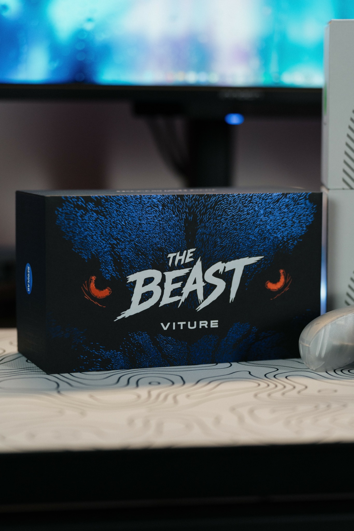 Box labeled 'The Beast' with a dark background and orange eyes, placed on a desk with a computer monitor behind.