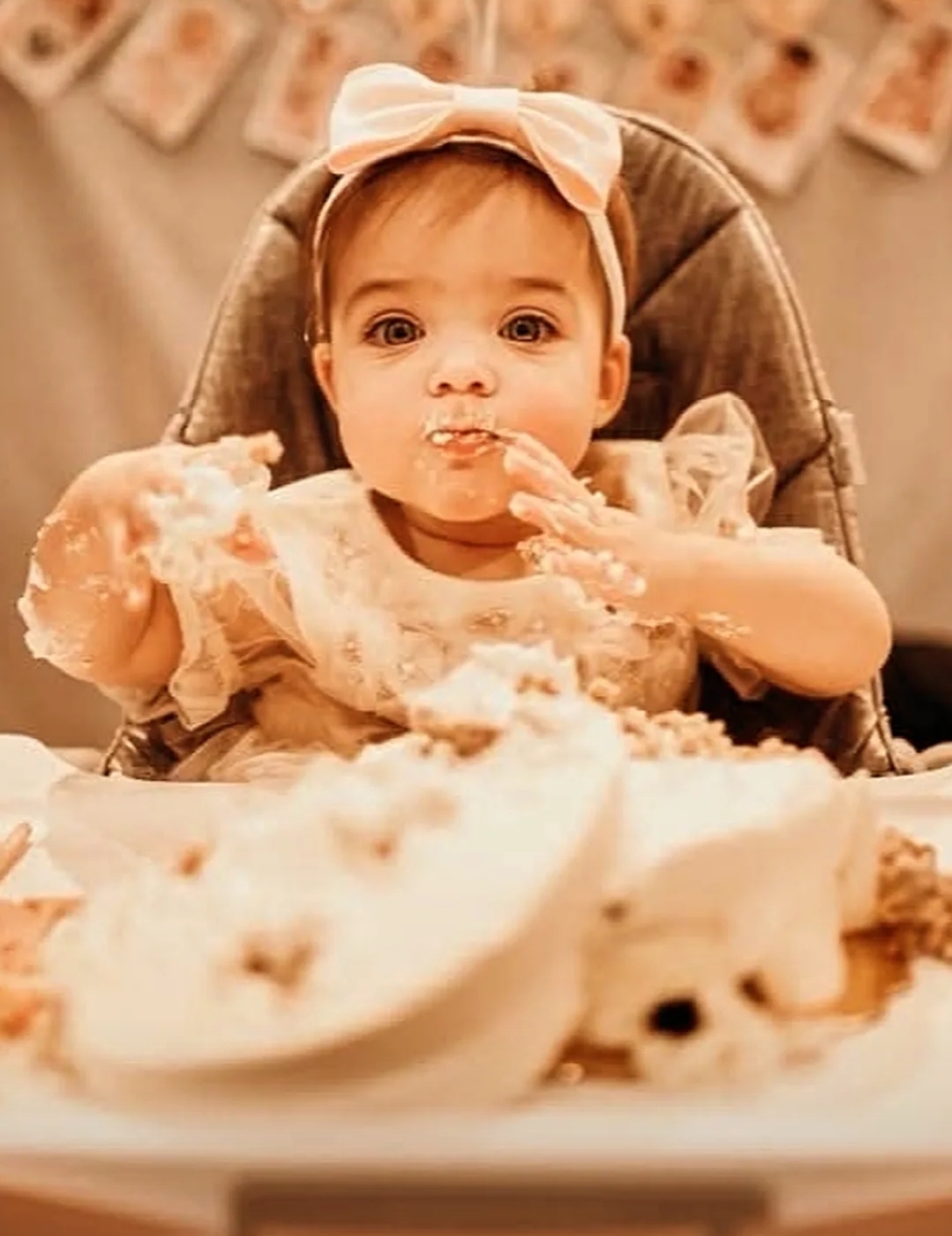 Baby girl with a bow on her head, sitting in a high chair, surrounded by cake and frosting, celebrating a birthday.
