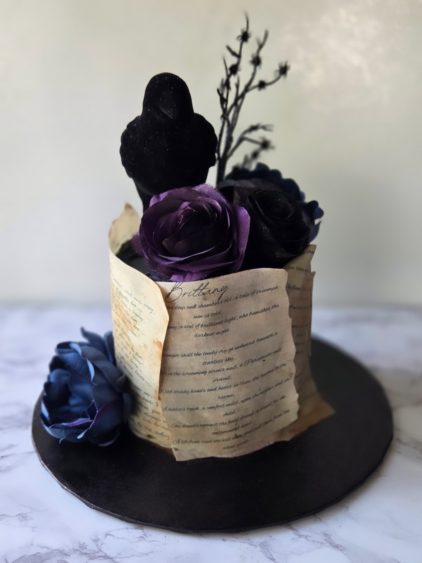 A decorative cake with purple and black flowers, a bird, and a paper scroll, on a black circular base.
