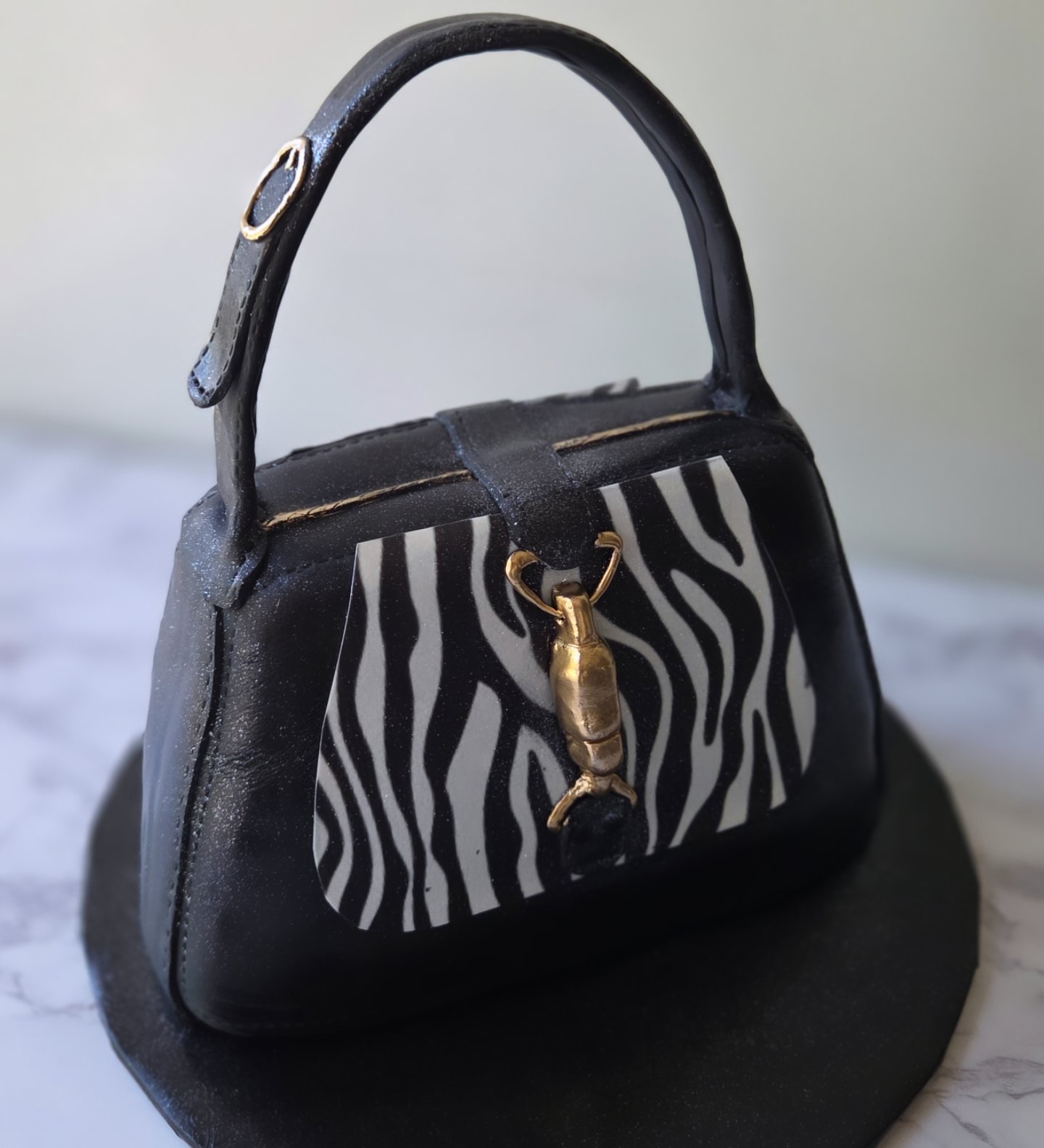 Small handbag with zebra pattern and gold animal charm, placed on a round black surface.