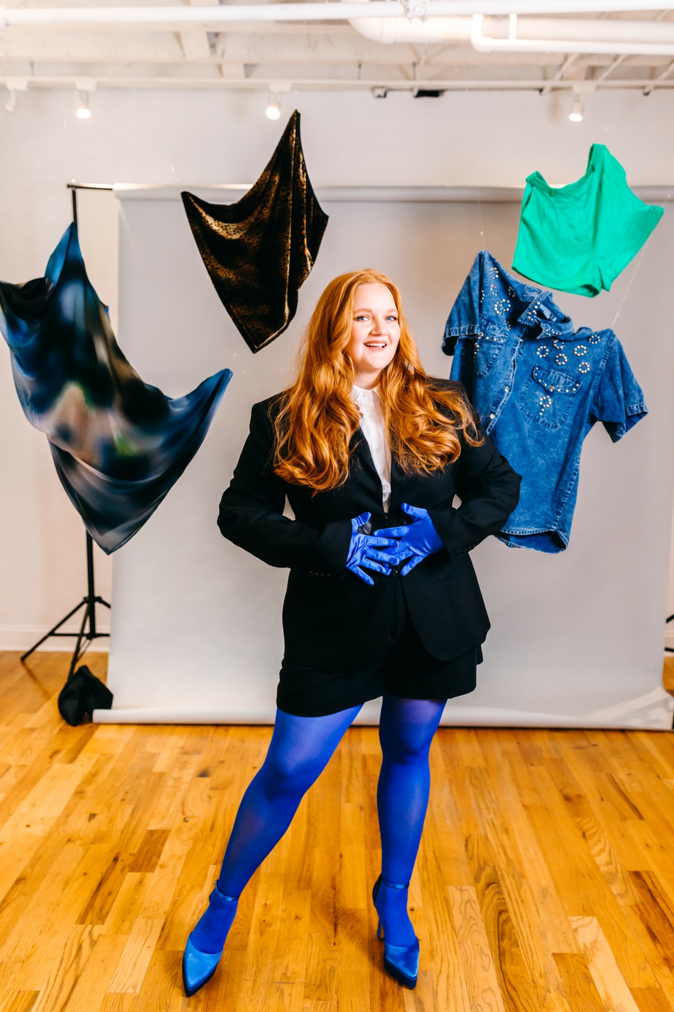 Woman with long red hair in a black coat and blue tights, smiling, surrounded by colorful fabric pieces hanging in a studio.
