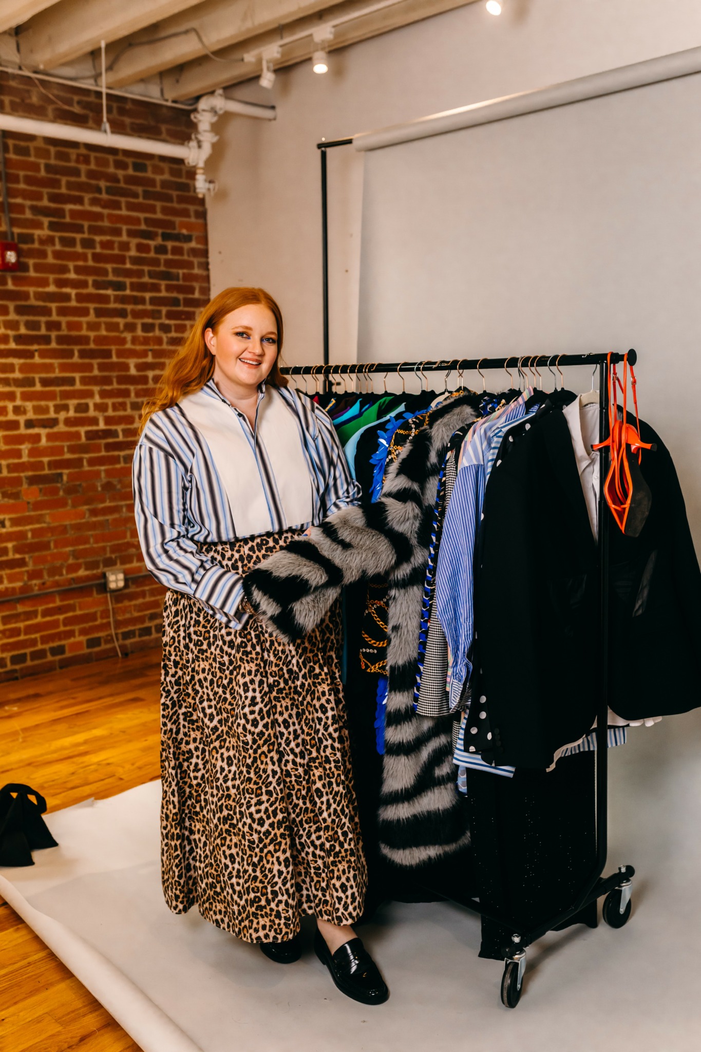 Woman with long hair wearing a striped shirt and leopard print skirt stands next to a clothing rack with various garments in a room with brick wall and whiteboard.