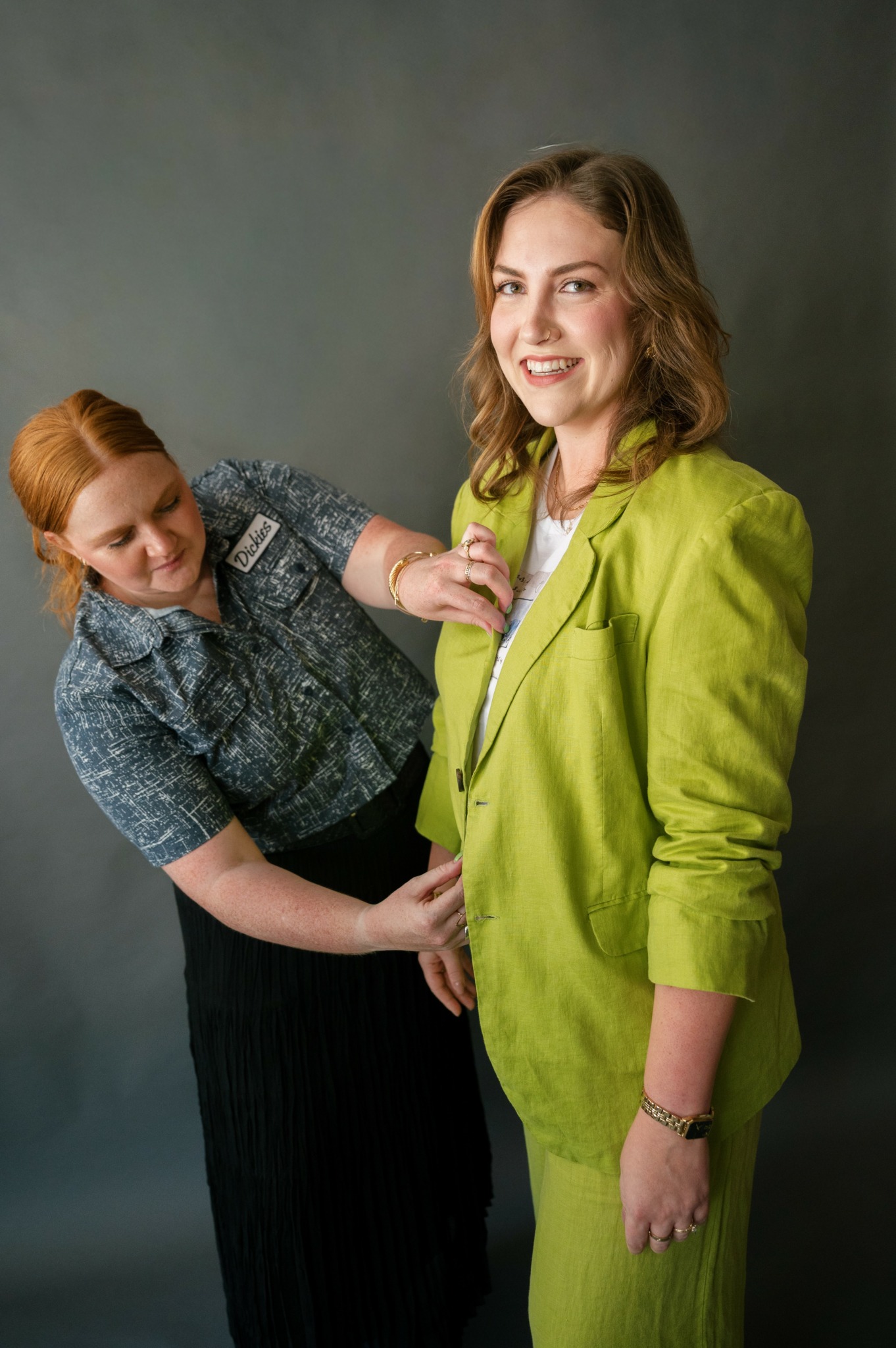 Two women, one in a green blazer and the other in a patterned shirt, smiling and interacting against a gray background.