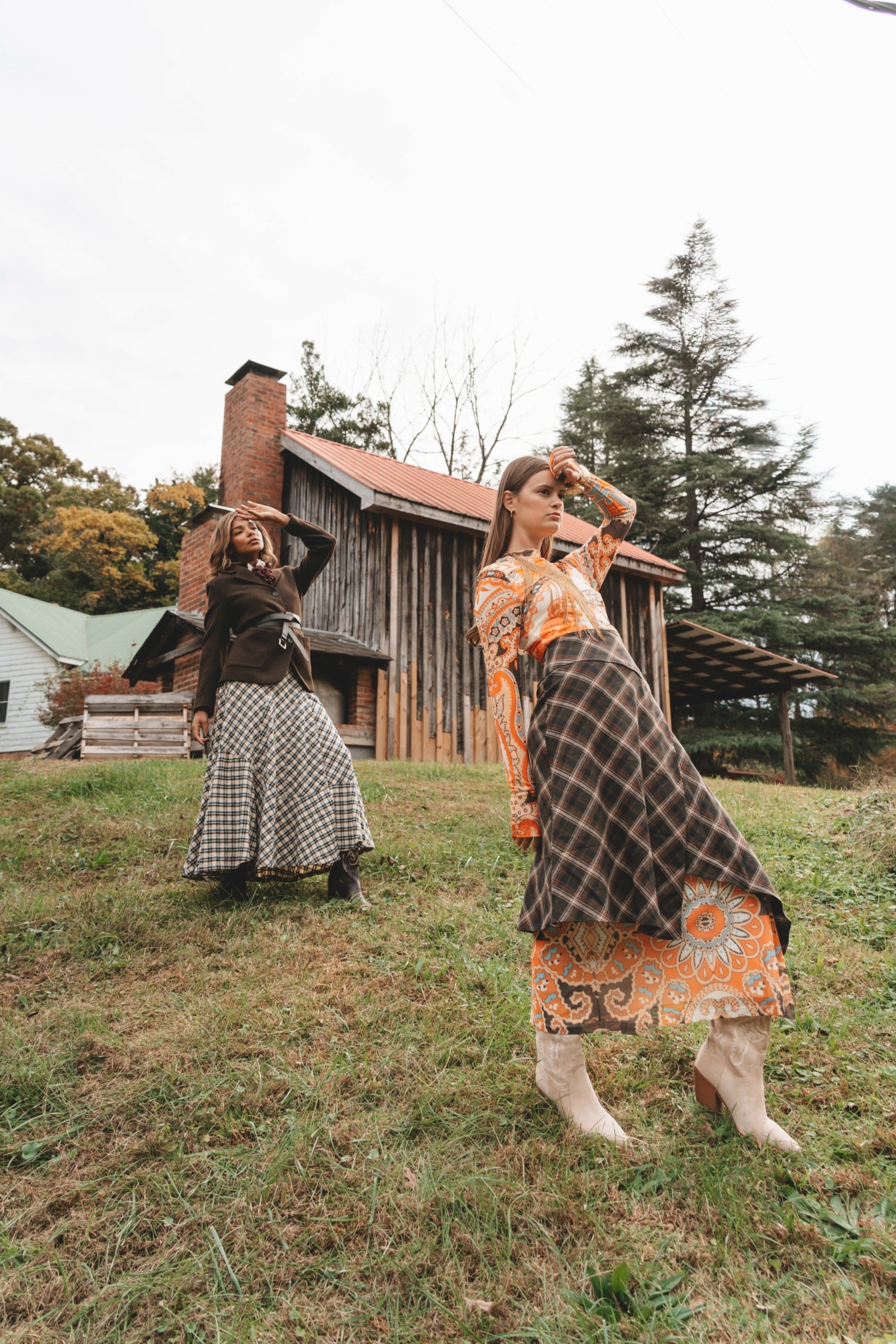 Two women in patterned skirts and tops stand outdoors on grass with trees and a wooden building in the background.