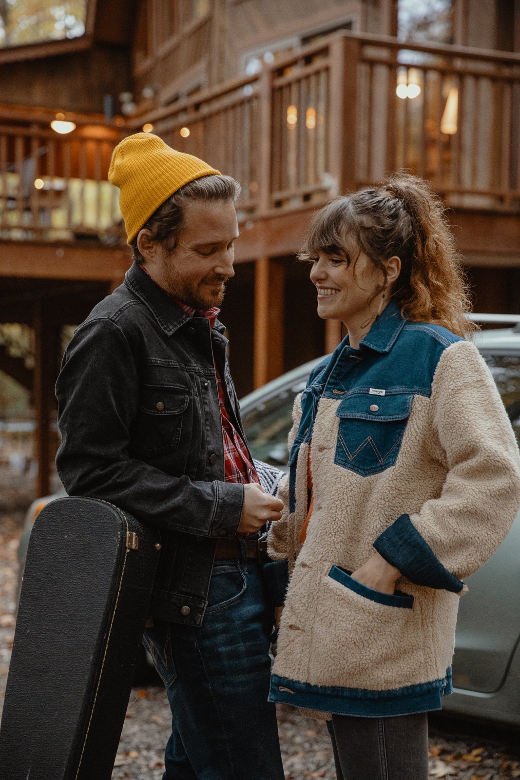 Two women smiling and talking outdoors near a car and wooden building, one wearing a yellow beanie and jacket, the other in a fleece jacket.