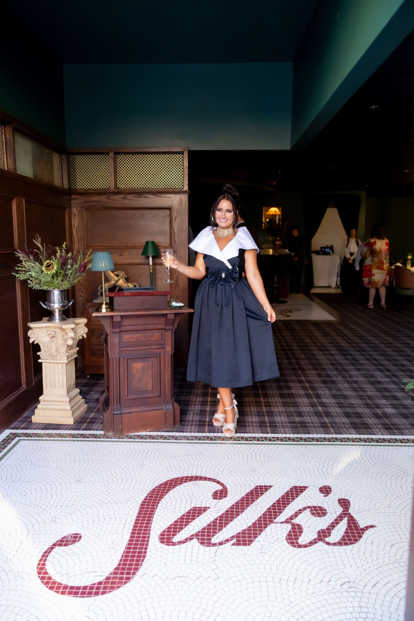 Woman in a navy blue dress standing indoors near a wooden table with a plant, in a decorated room.