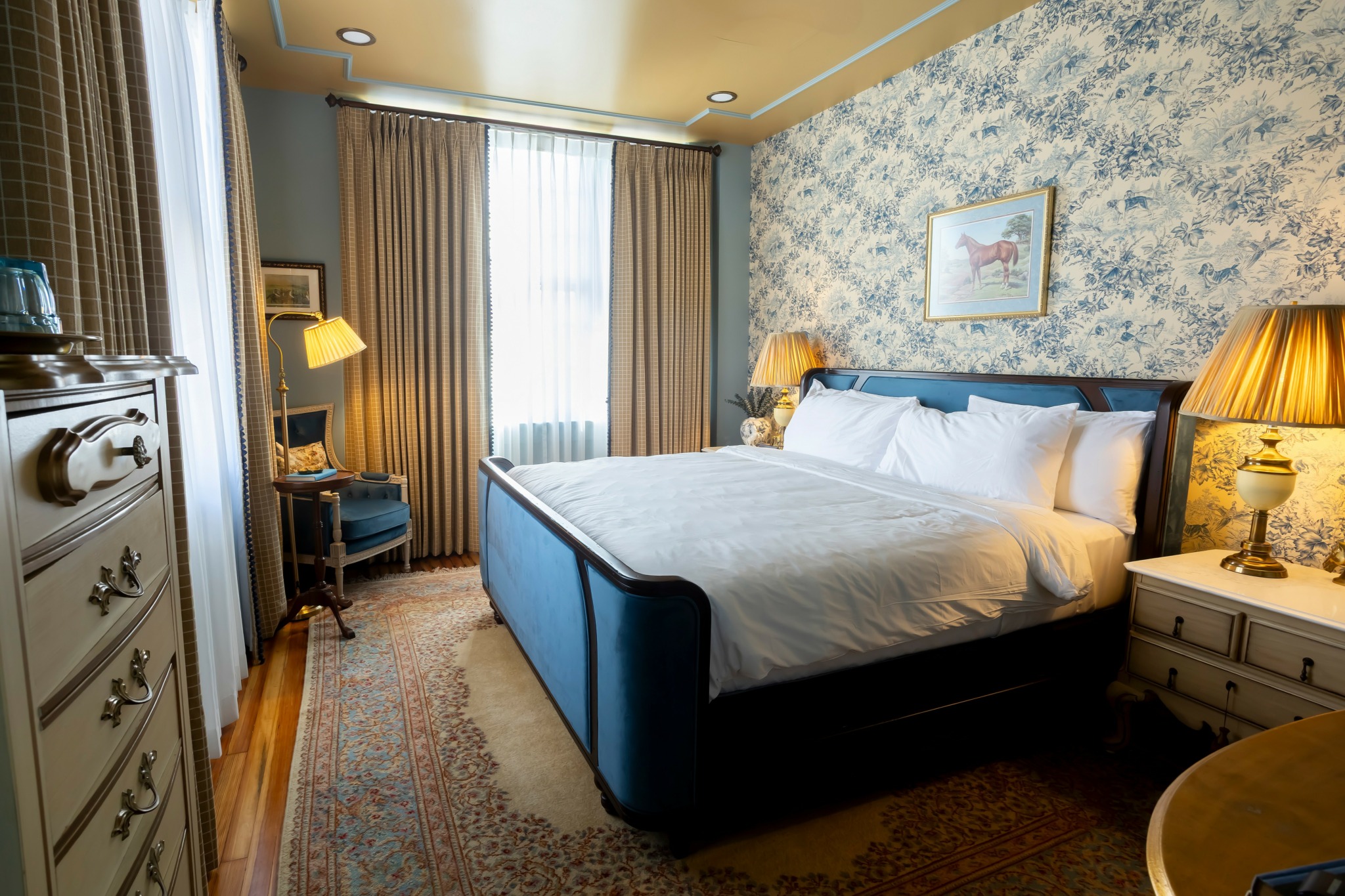 Bedroom with a large bed, nightstands, lamps, window with curtains, and a dresser, decorated with floral wallpaper.
