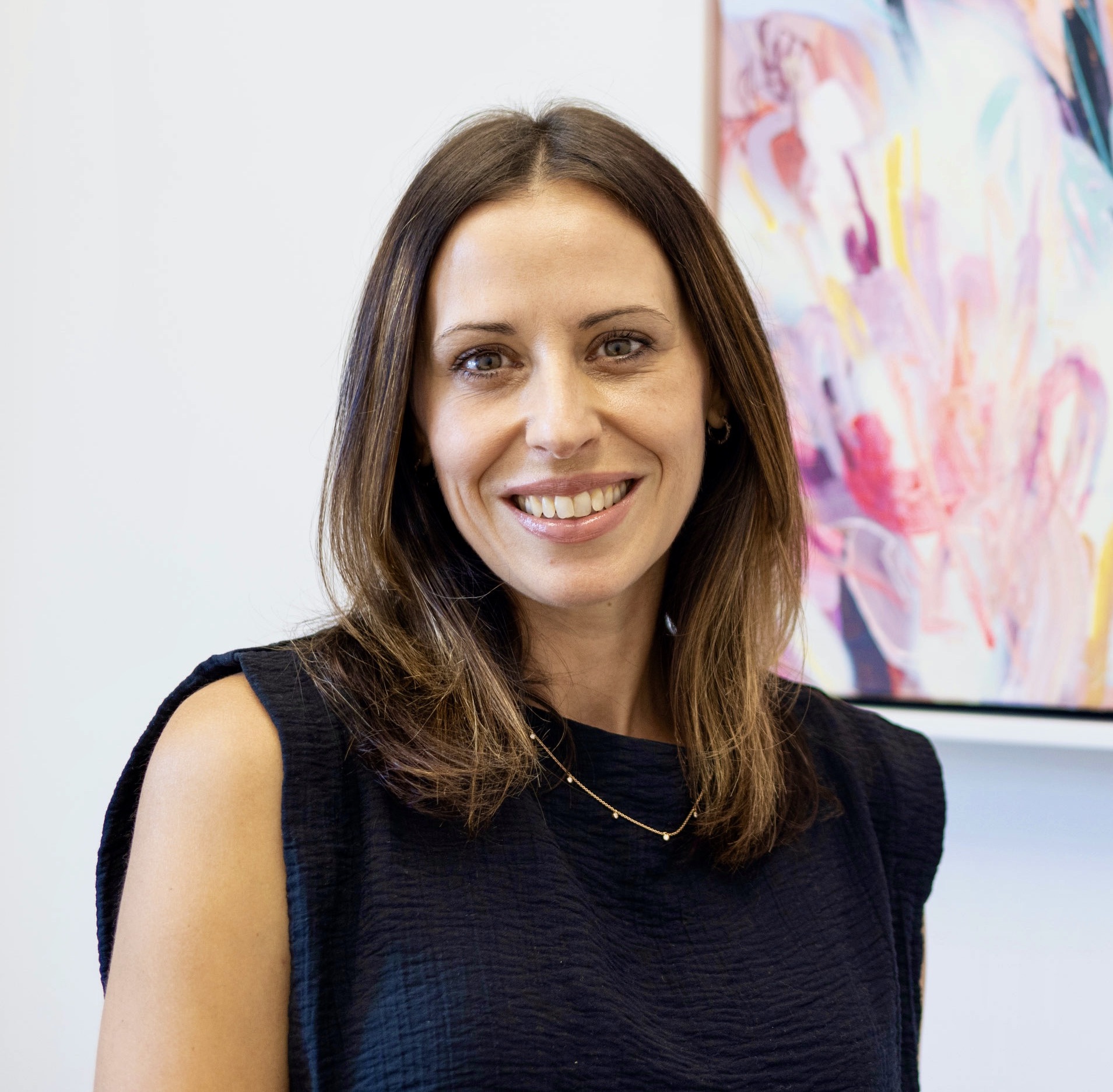 Woman with shoulder-length brown hair smiling, wearing a black sleeveless top, standing in front of colorful abstract art.
