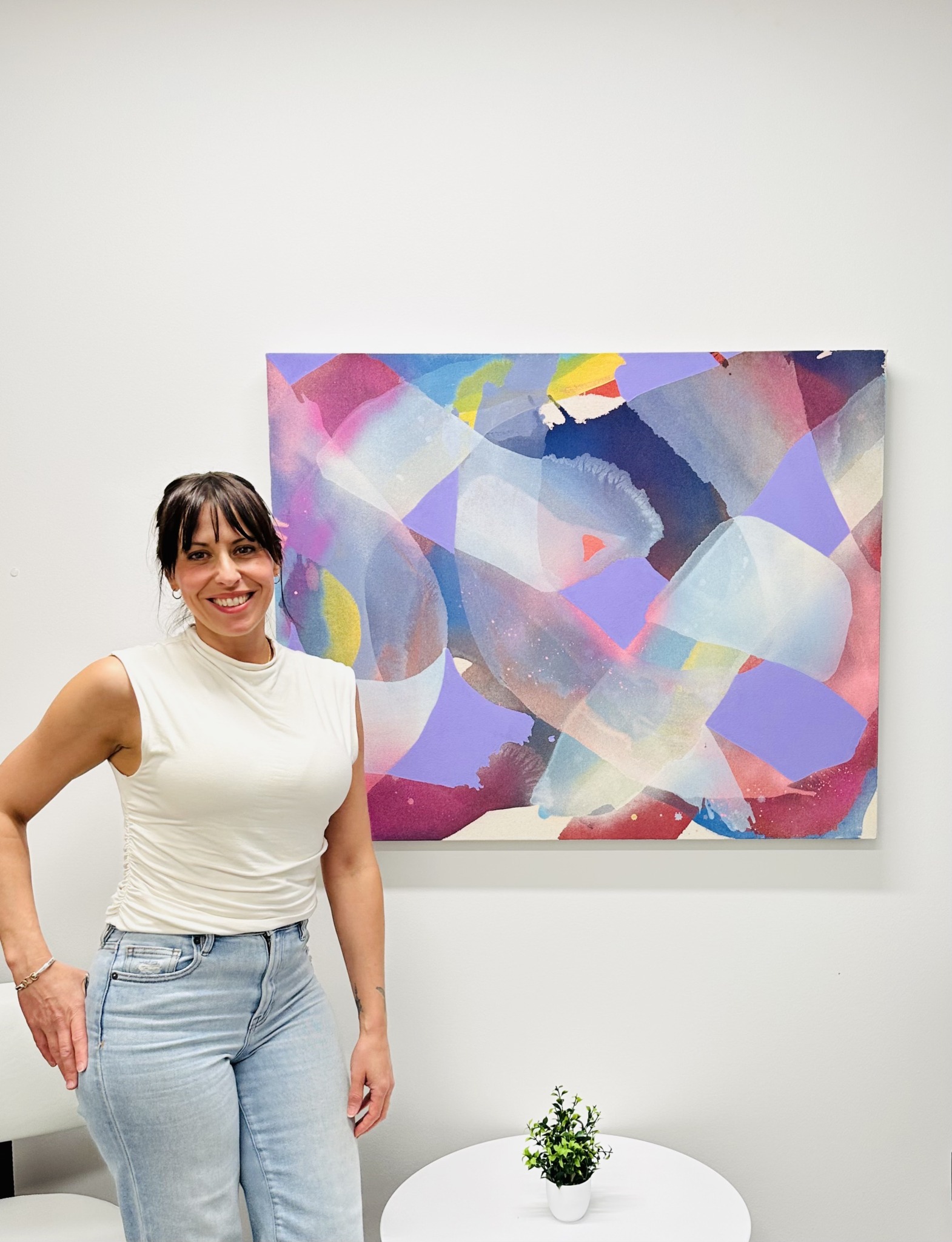 Woman with dark hair in a white sleeveless top and light jeans standing next to colorful abstract painting on white wall, smiling.