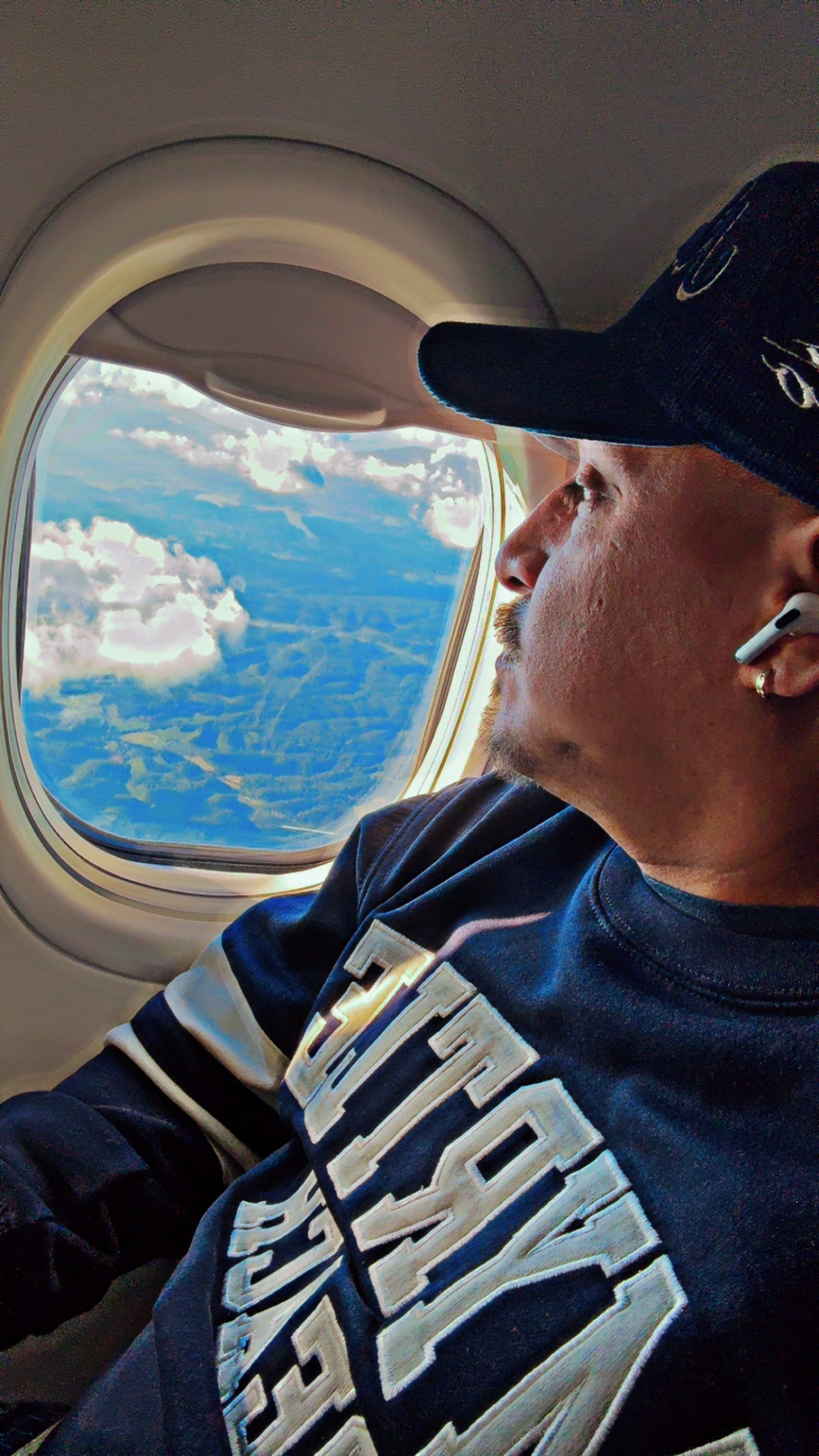 Person wearing a cap and earbud looking out airplane window at clouds and landscape.