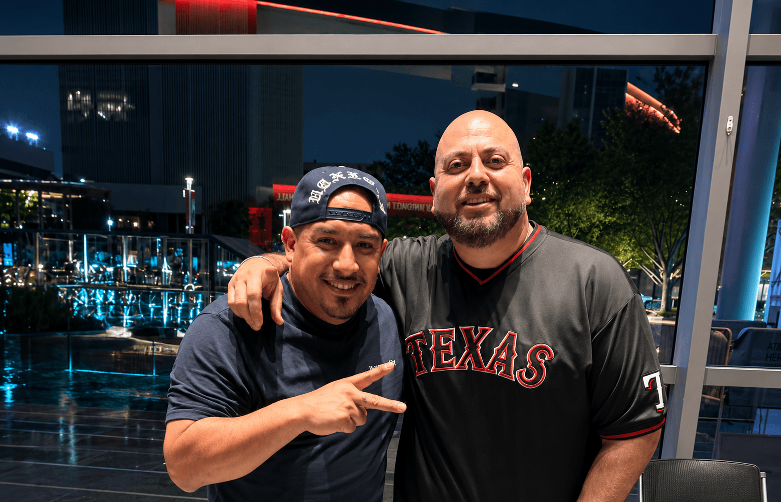 Two men standing indoors at night, one wearing a cap and pointing, the other smiling with arm around shoulder.