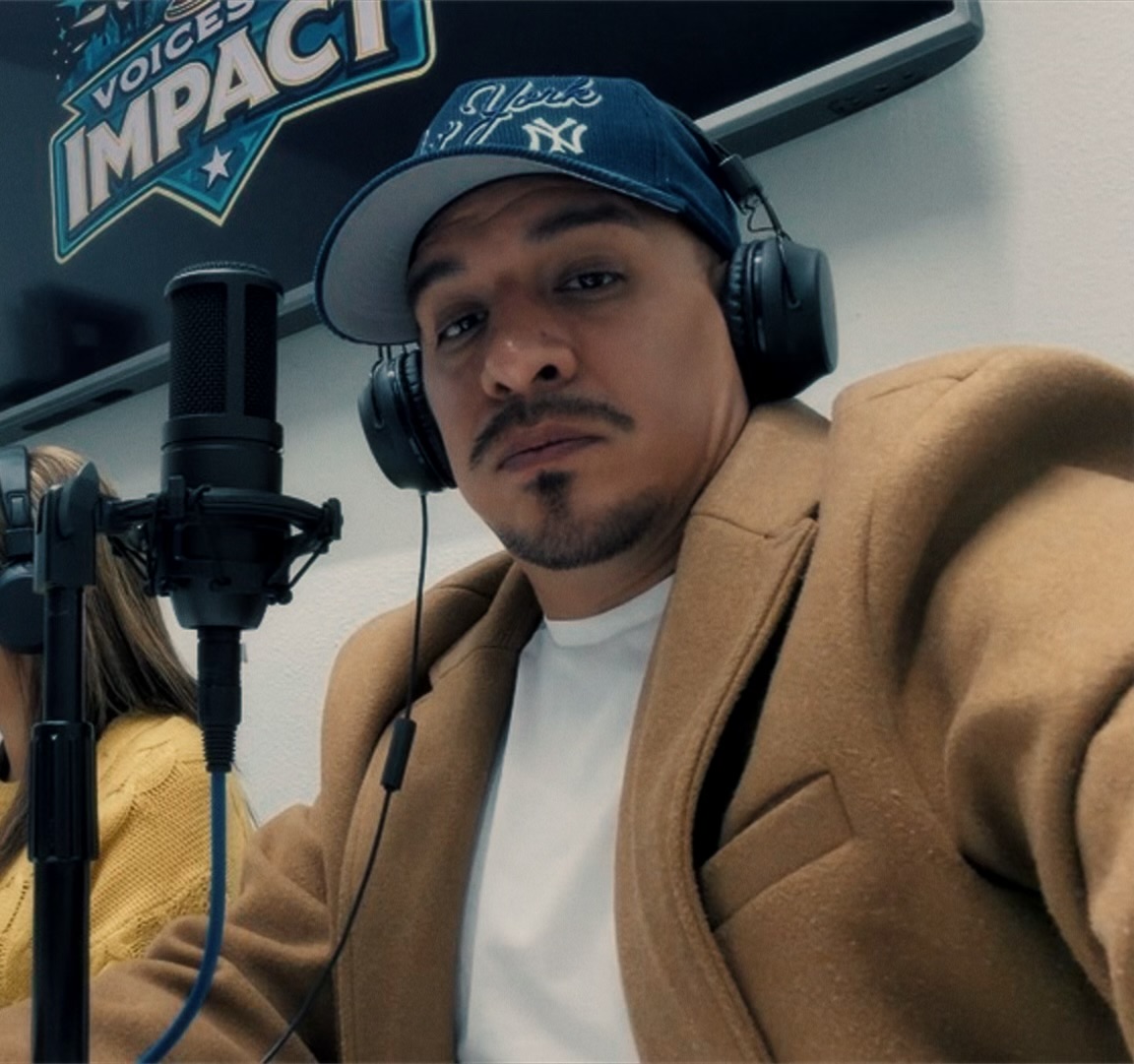 Man wearing a blue cap and headphones, sitting in front of a microphone, with a sign in the background.
