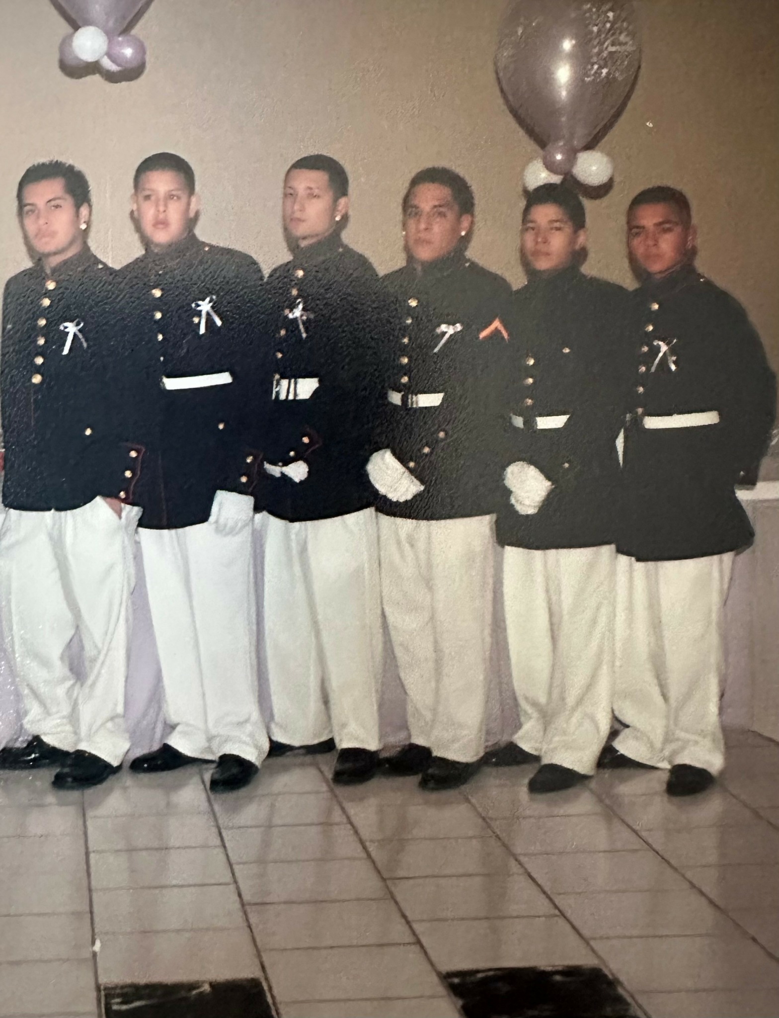 Six young men in formal uniforms standing indoors, with balloons overhead, facing forward, hands at sides, tiled floor beneath.