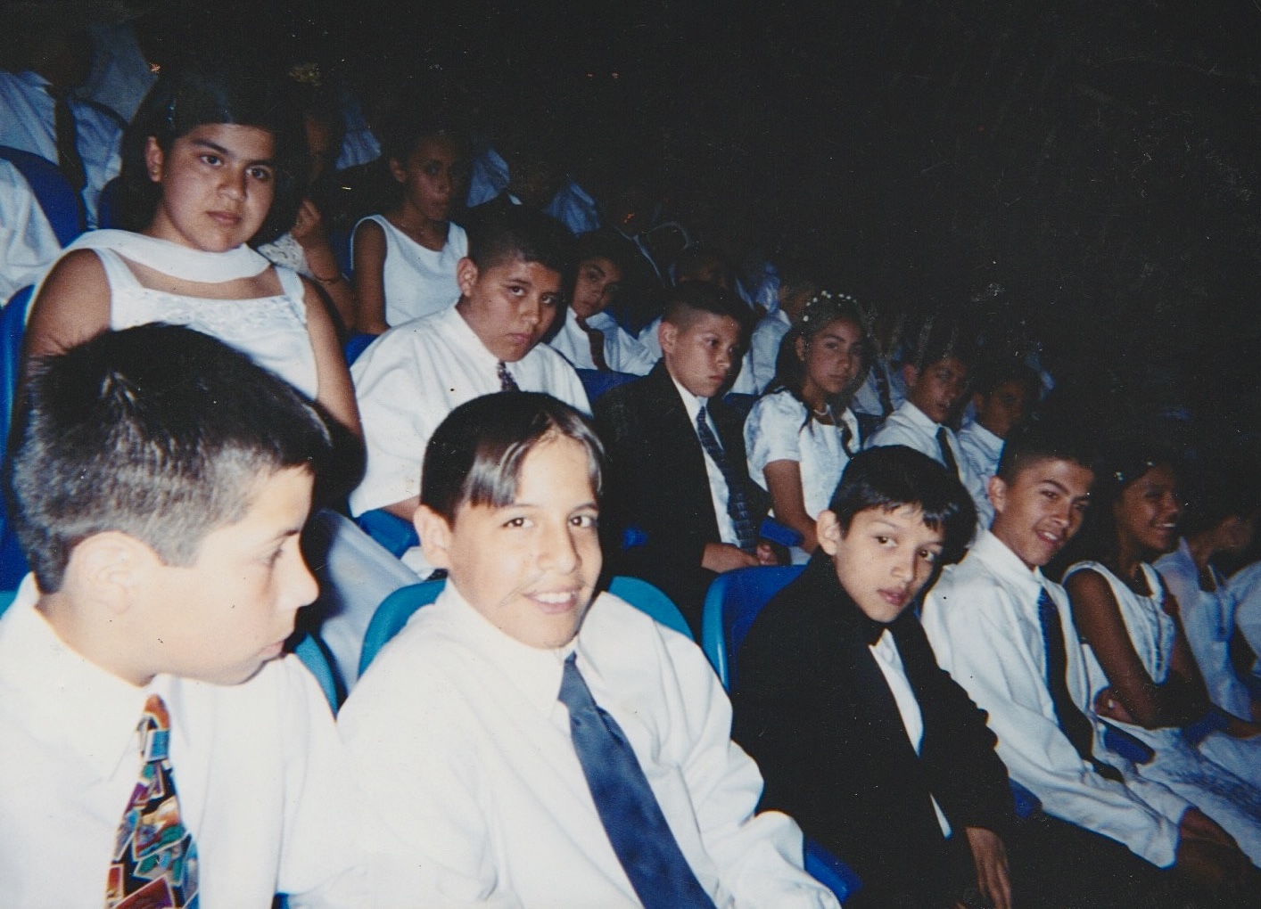Group of children sitting in a dark auditorium, some smiling, wearing formal clothes, some in white shirts and ties.