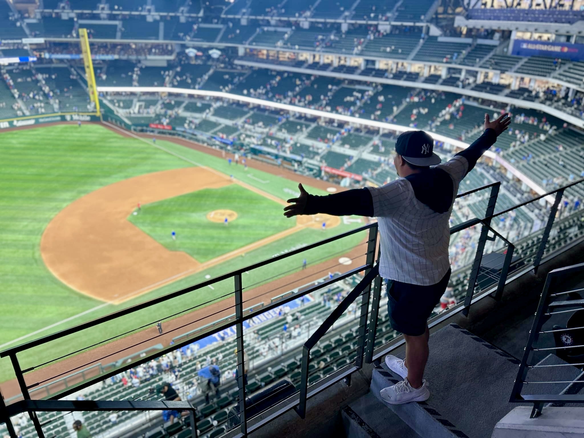 Child standing on stadium balcony, arms outstretched, overlooking baseball field with empty seats.