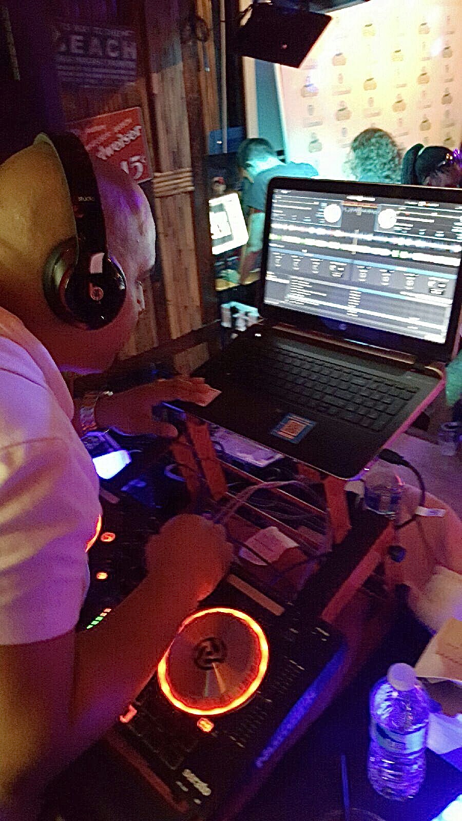 Person DJing with headphones, using a laptop and DJ equipment with colorful lights, in a dimly lit environment.