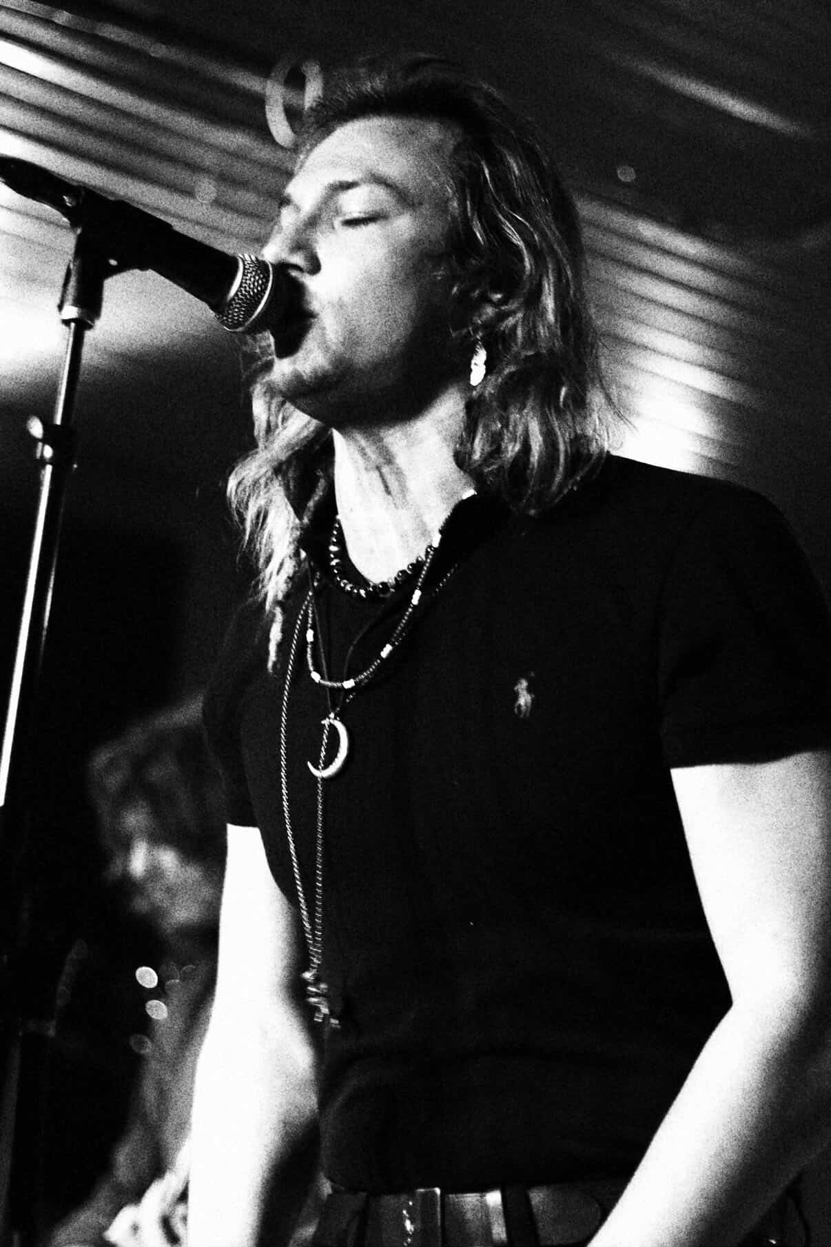 Person singing into microphone with eyes closed, long hair, wearing dark clothing and layered necklaces, stage background.