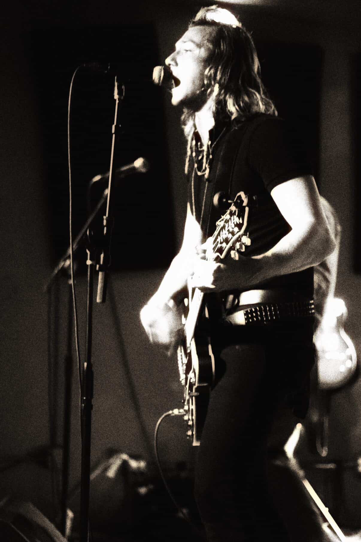 Musician singing into microphone while playing guitar on stage, with a dark background.