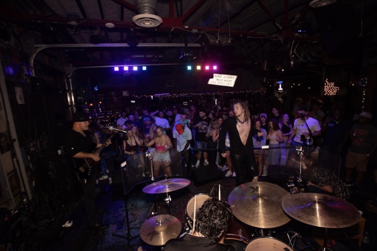 Band performing on stage with a crowd in a dark venue, colorful lights overhead, and musical instruments visible.