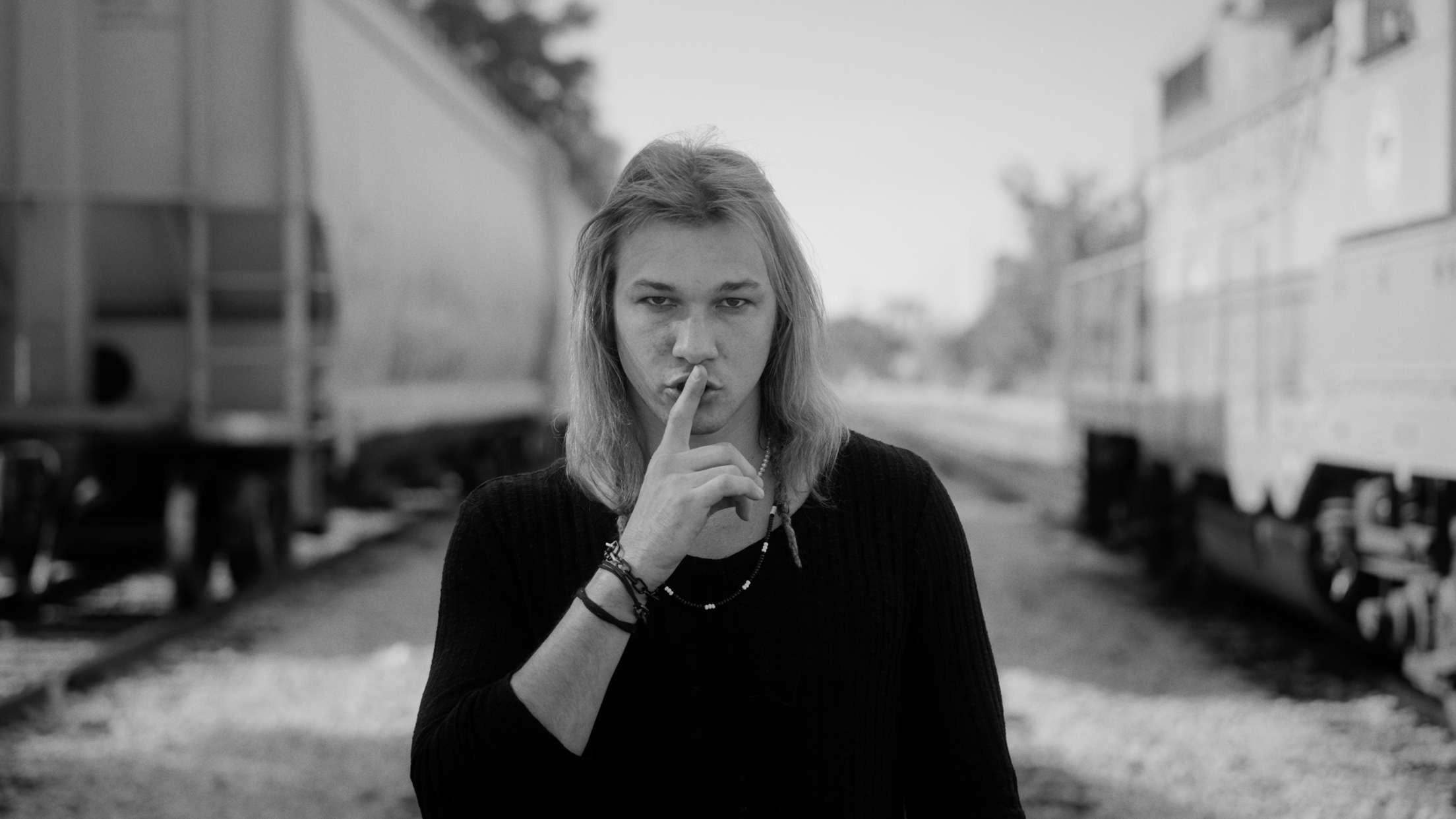 Young woman with long hair making a peace sign near her face, standing between train cars, in black and white.