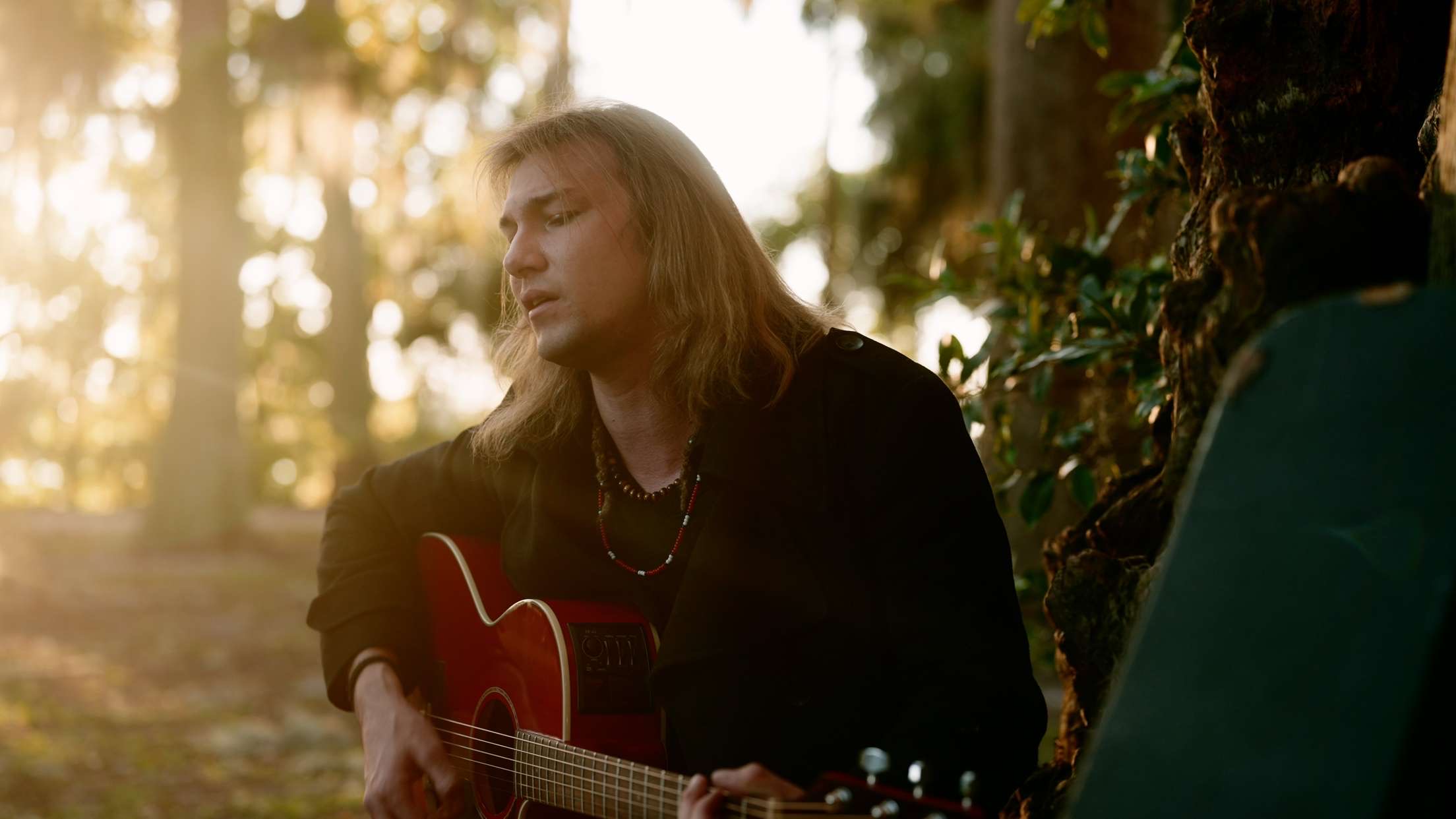 Person with long hair playing guitar outdoors in a forest setting, sunlight filtering through trees.