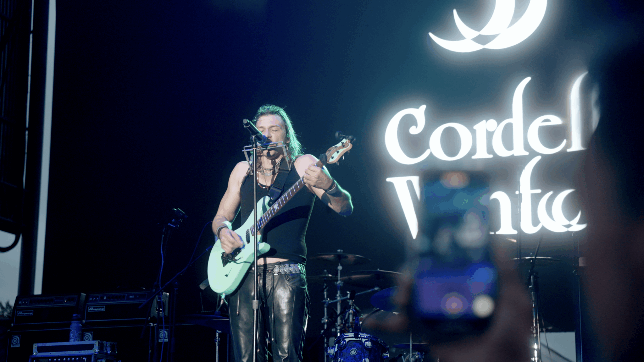 Musician with green hair playing guitar on stage with a large illuminated sign behind her.